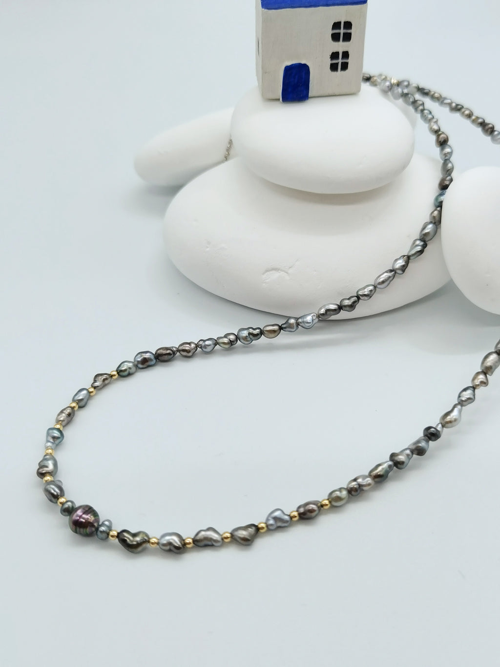 HEART PEARL NECKLACE WITH GOLD BEADS arranged in a stylish curve flowing from the little house on top of some white stones in the background, showing off the multiple coloured strand with brilliance contrast from the gold beads