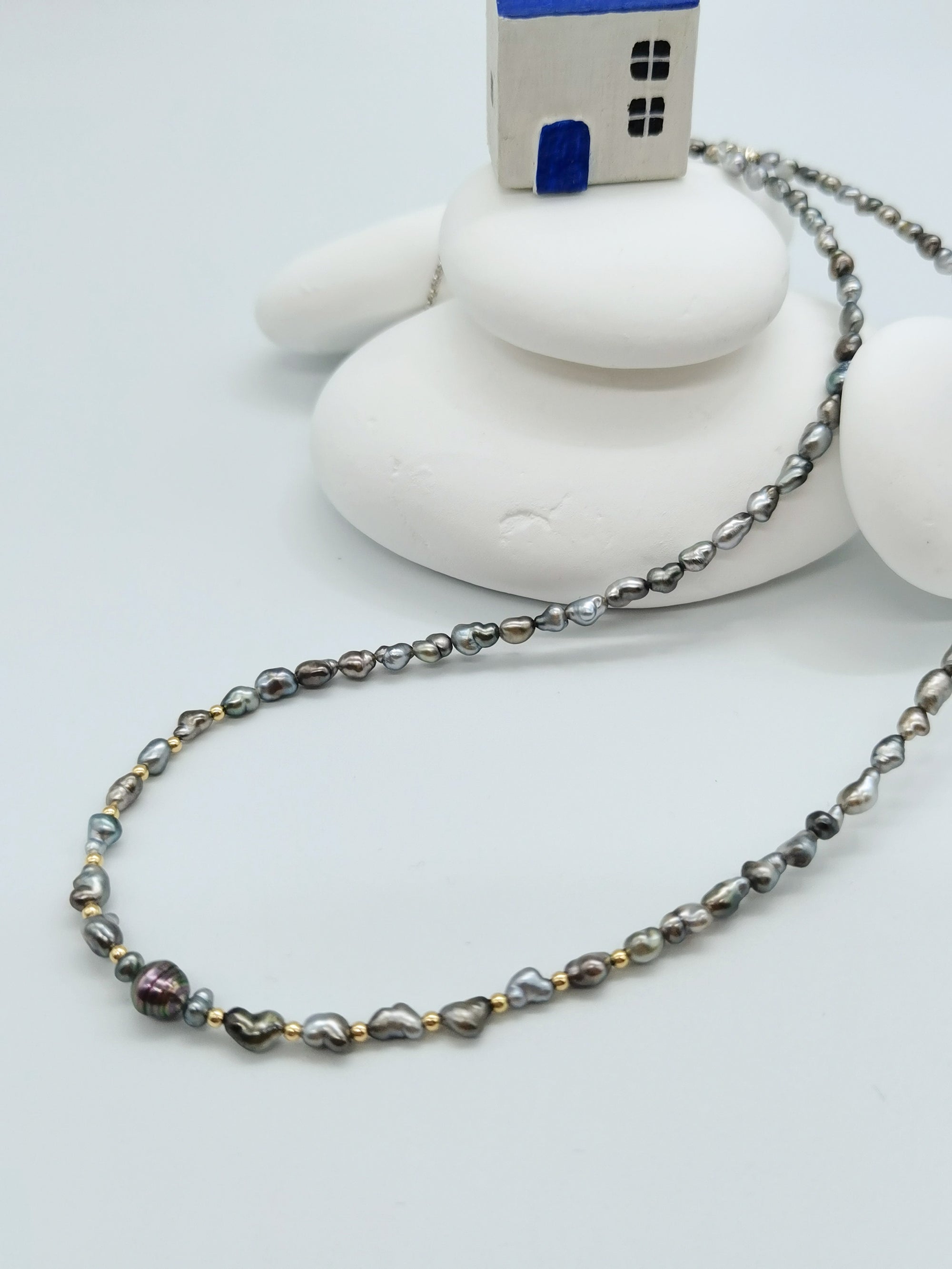HEART PEARL NECKLACE WITH GOLD BEADS arranged in a stylish curve flowing from the little house on top of some white stones in the background, showing off the multiple coloured strand with brilliance contrast from the gold beads