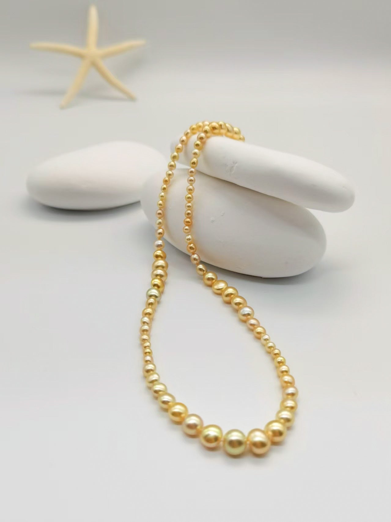 Full-length view of golden pearl strand necklace elegantly draped over white smooth stones with starfish accent, showcasing the complete graduated design from smallest to largest pearls