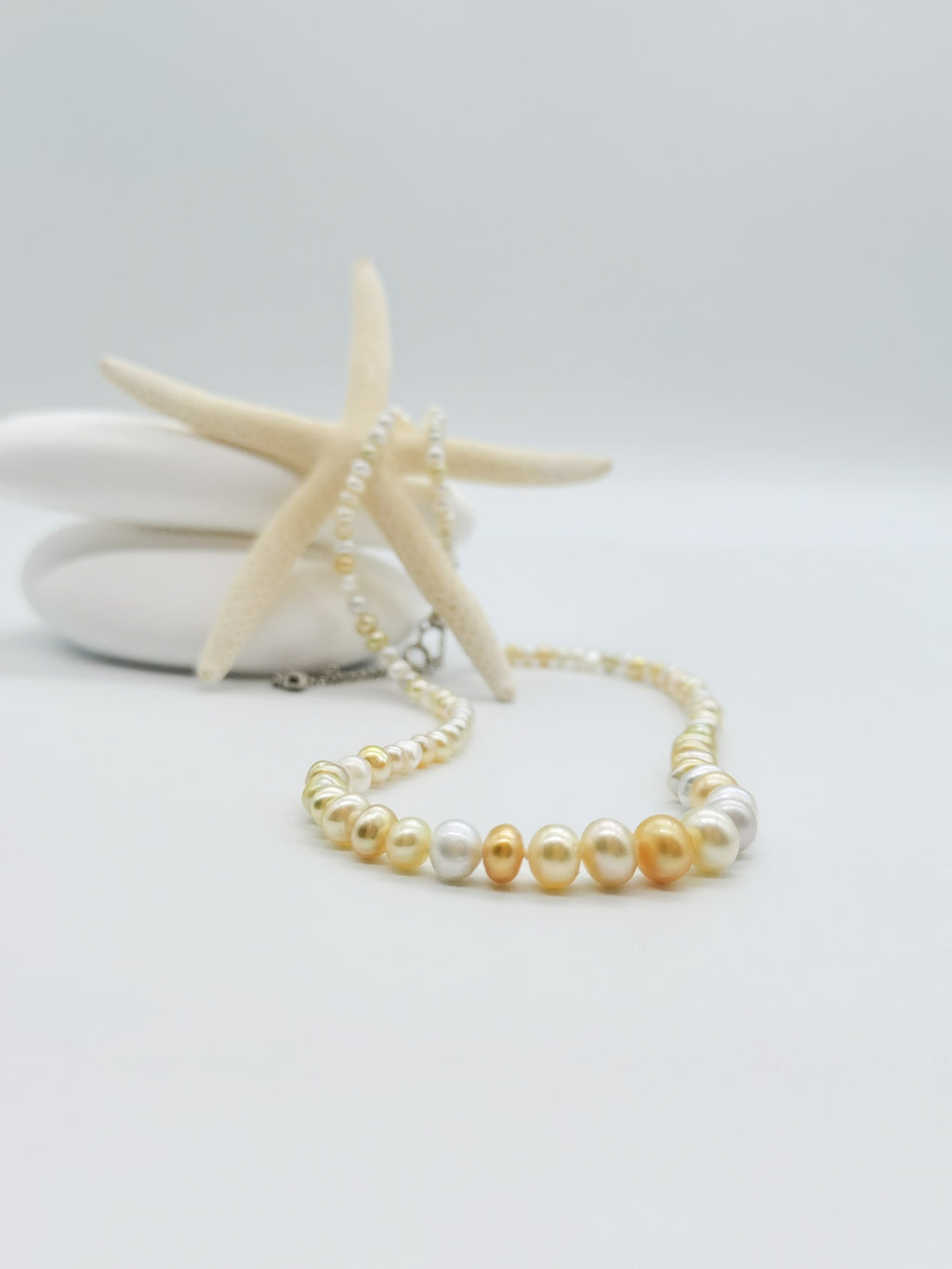 Close-up detail of white and cream pearl necklace draped over starfish against white stones, highlighting the lustrous graduated pearls and silver clasp