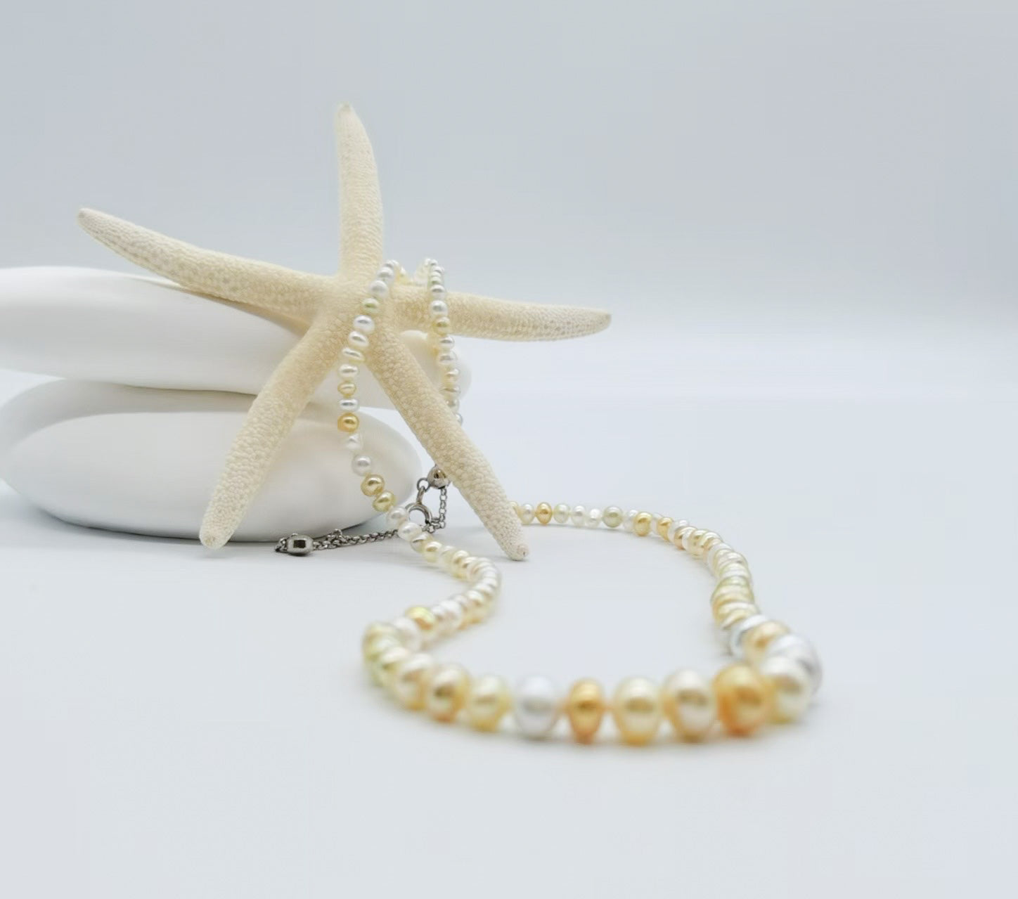 White and cream pearl strand necklace elegantly draped around starfish on white stones, showcasing the graduated pearl design with mixed white, cream and golden tones