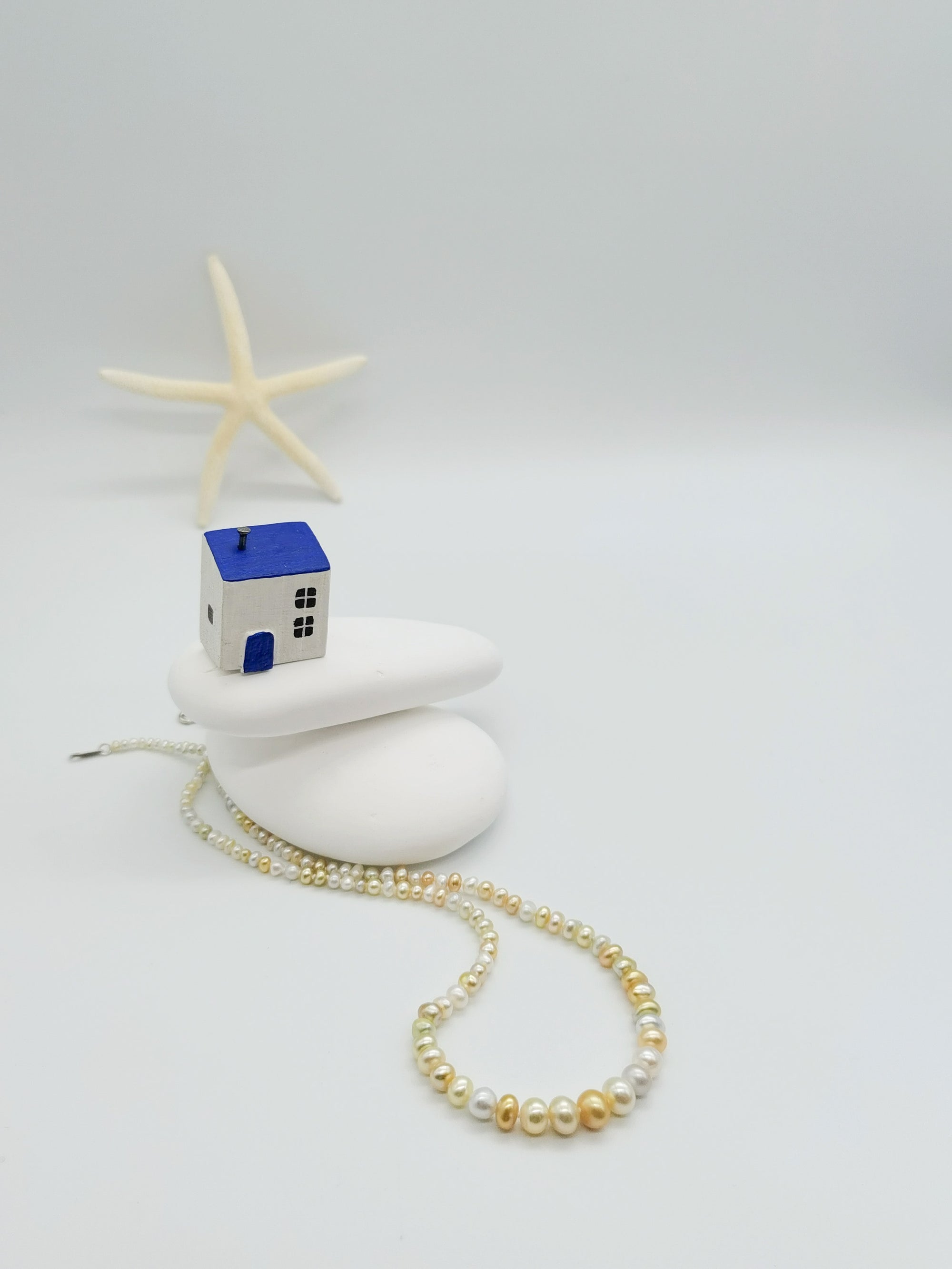 White pearl strand necklace artistically arranged on white stones with decorative blue house ornament and starfish in coastal-themed styling