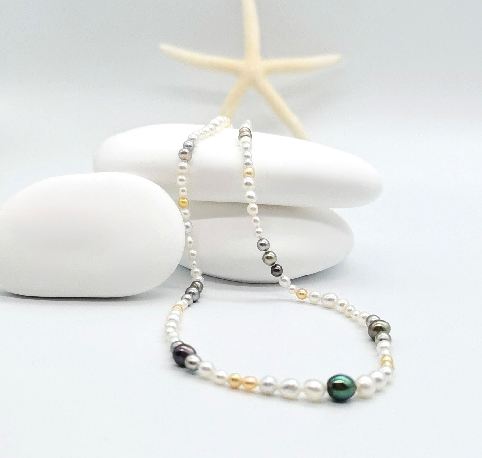 Multi-coloured pearl strand necklace elegantly draped in white shell with starfish accent, showcasing the beautiful mix of white, golden, silver, and dark green pearls in coastal styling