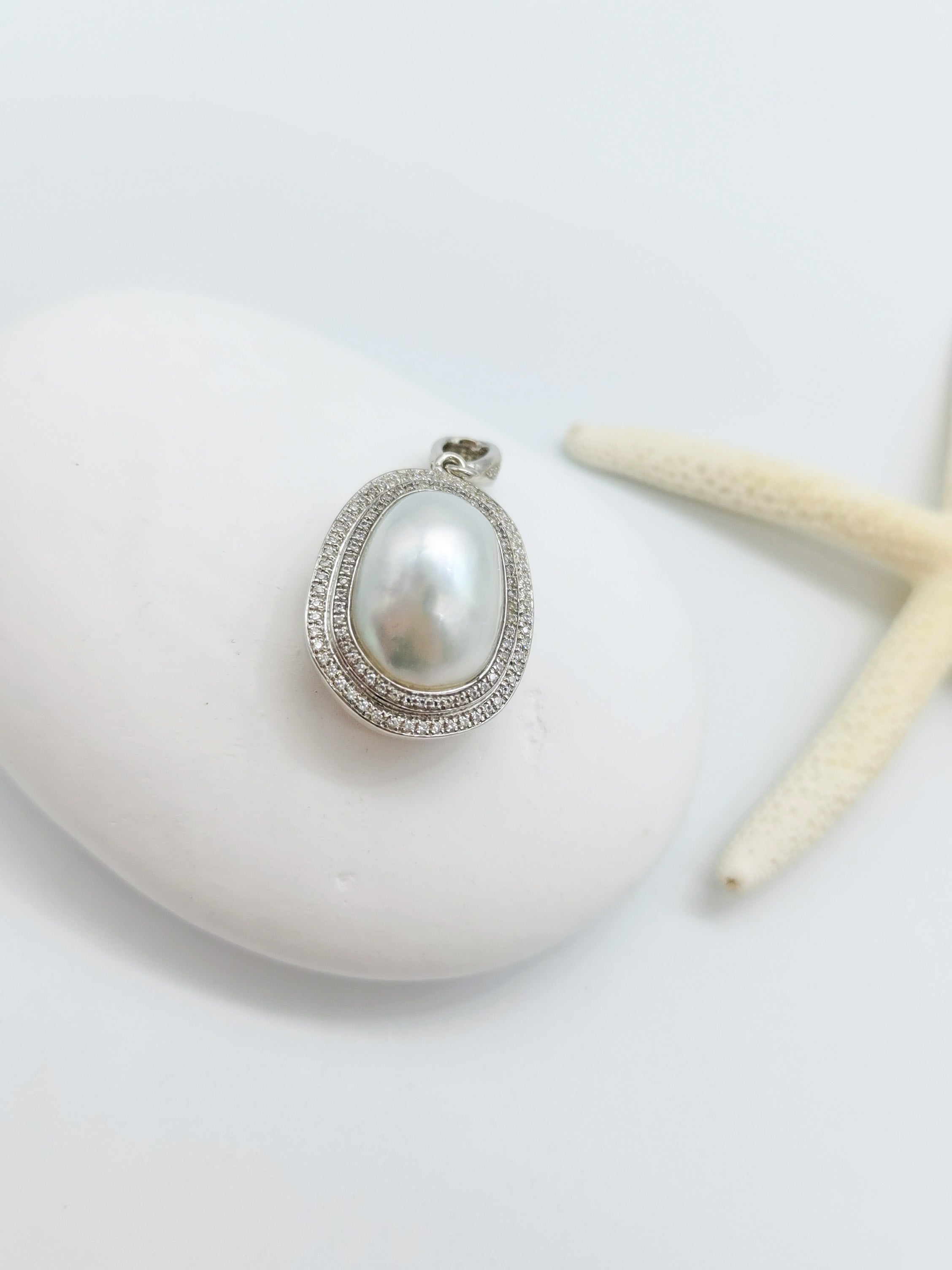 Detailed view of oval white pearl pendant with sparkling crystal surround and silver setting, artistically positioned on white stone with starfish accent