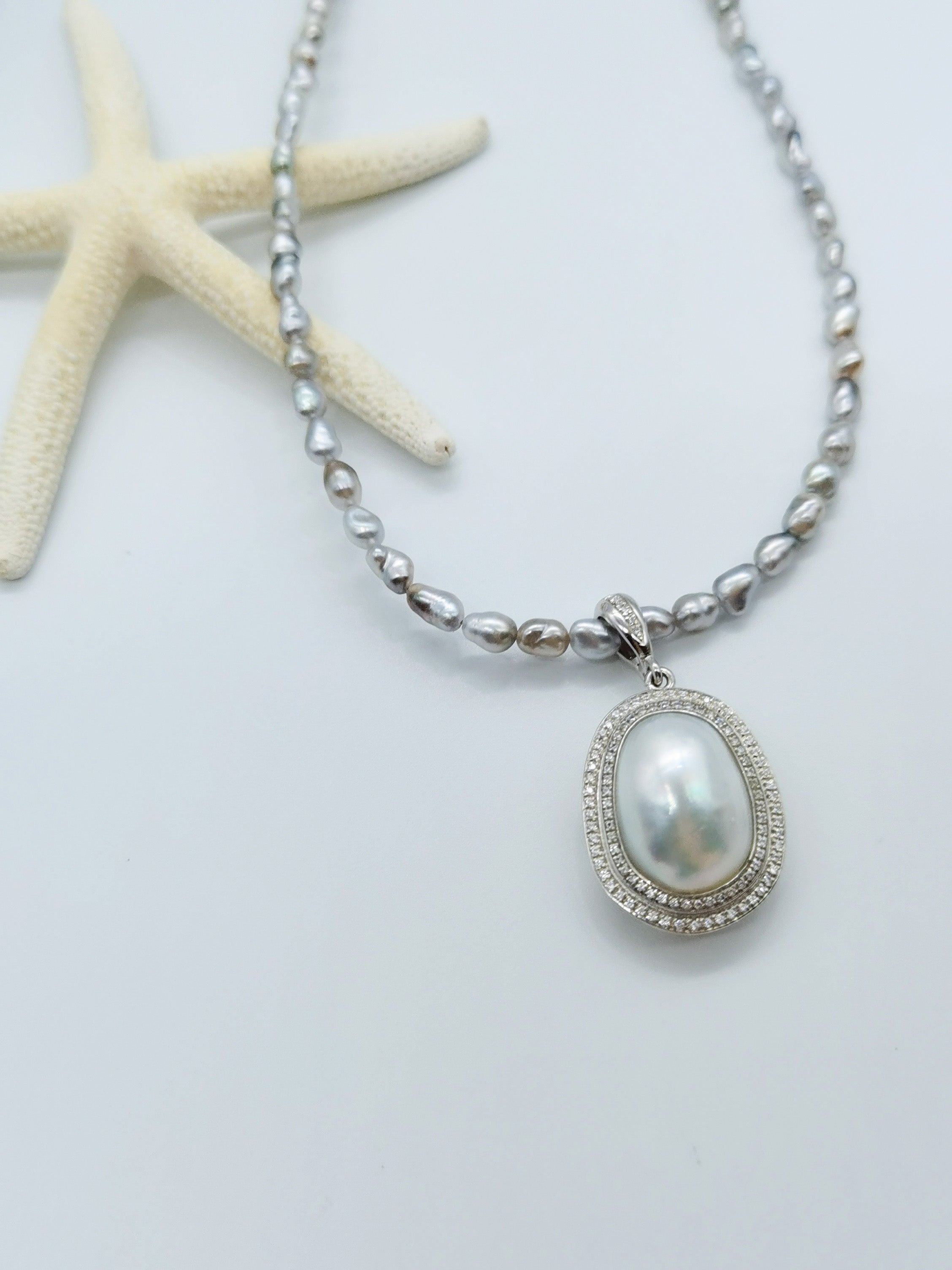 Grey baroque pearl necklace with oval white pearl pendant surrounded by crystal halo, displayed with white starfish on clean white background