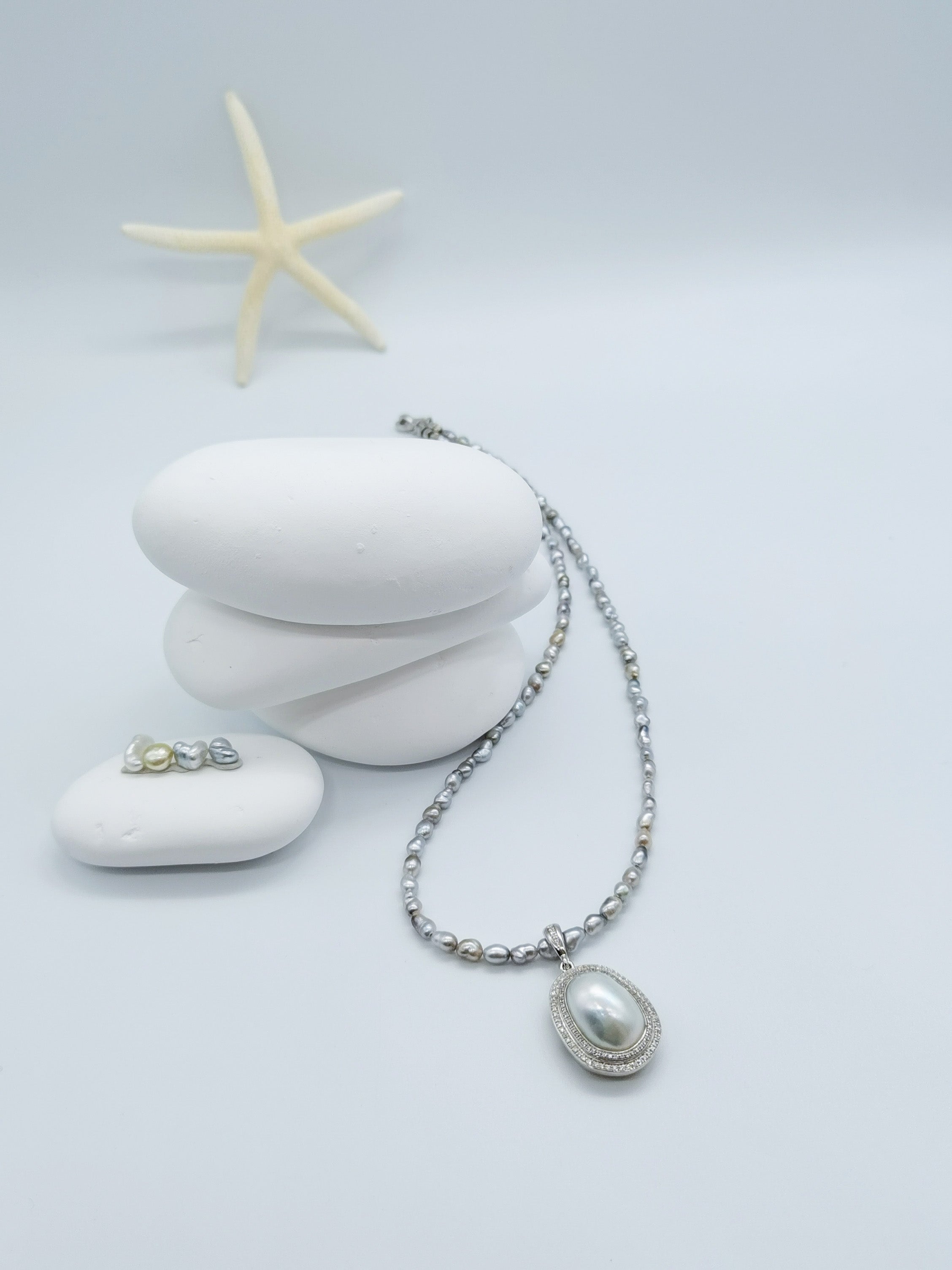 Grey baroque pearl necklace with white oval pearl and crystal halo pendant, elegantly draped over white stones with starfish accent in coastal styling