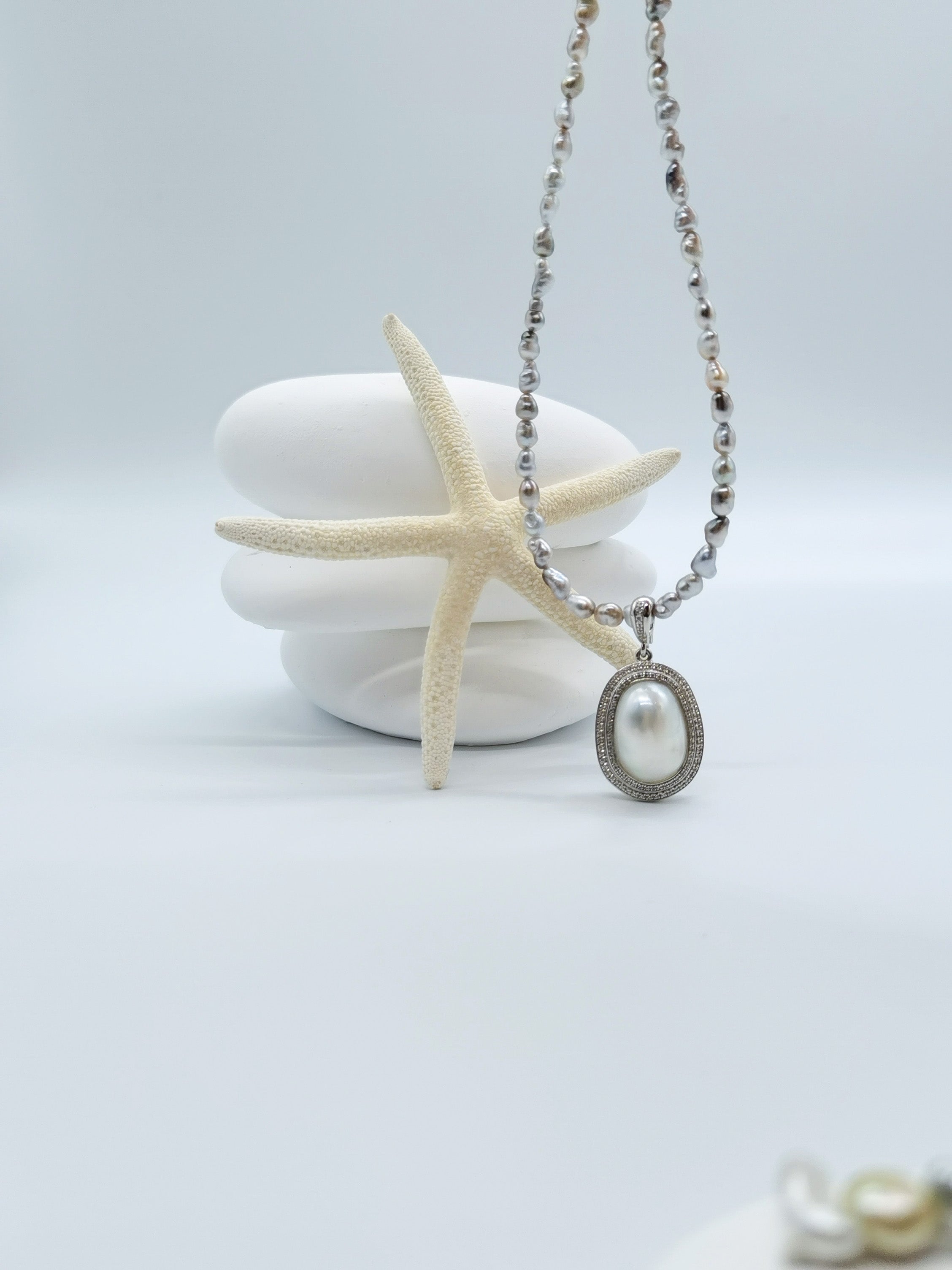 Grey baroque pearl strand necklace featuring oval white pearl pendant with sparkling crystal surround, artistically arranged with starfish on stacked white stones