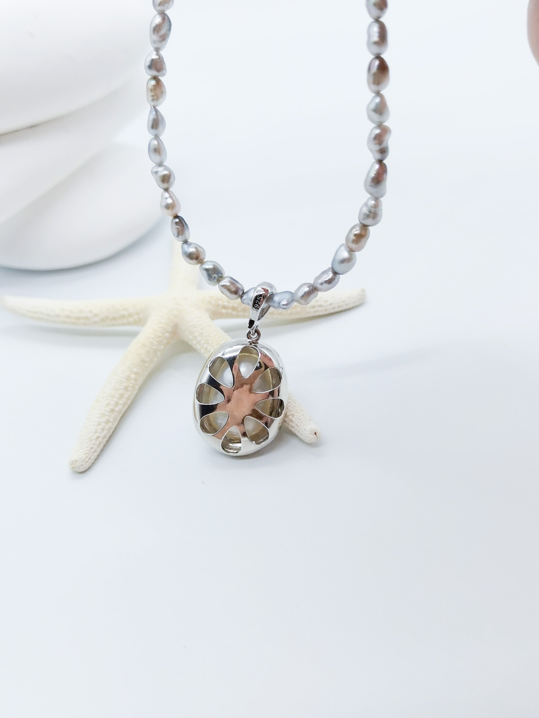 White oval pearl pendant with crystal halo displayed on smooth white stone, showcasing the intricate silver setting and open back setting with starfish backdrop