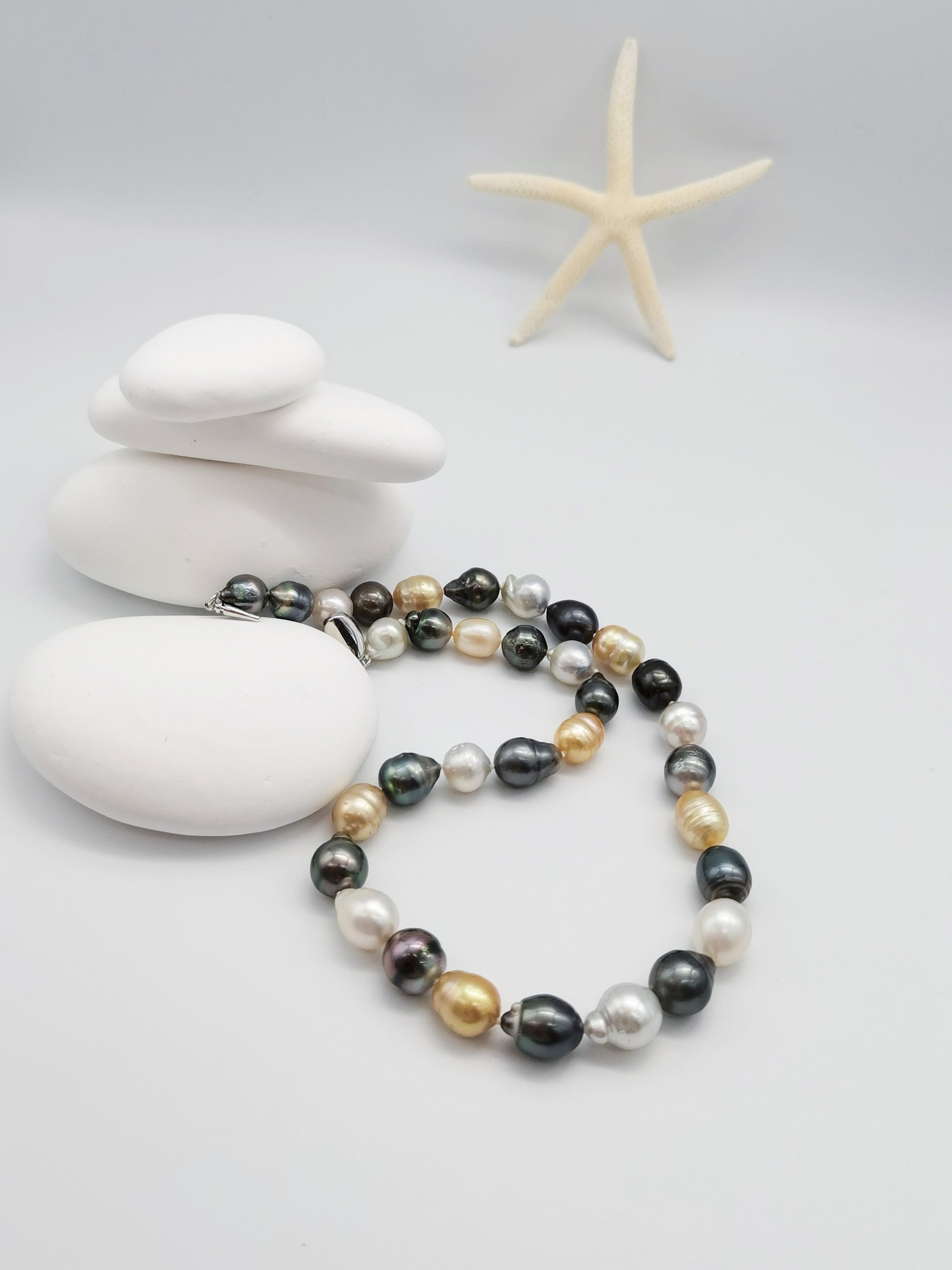 Multicoloured baroque pearl necklace with white, golden, dark green and black pearls artistically arranged on stacked white stones with starfish in zen-like composition