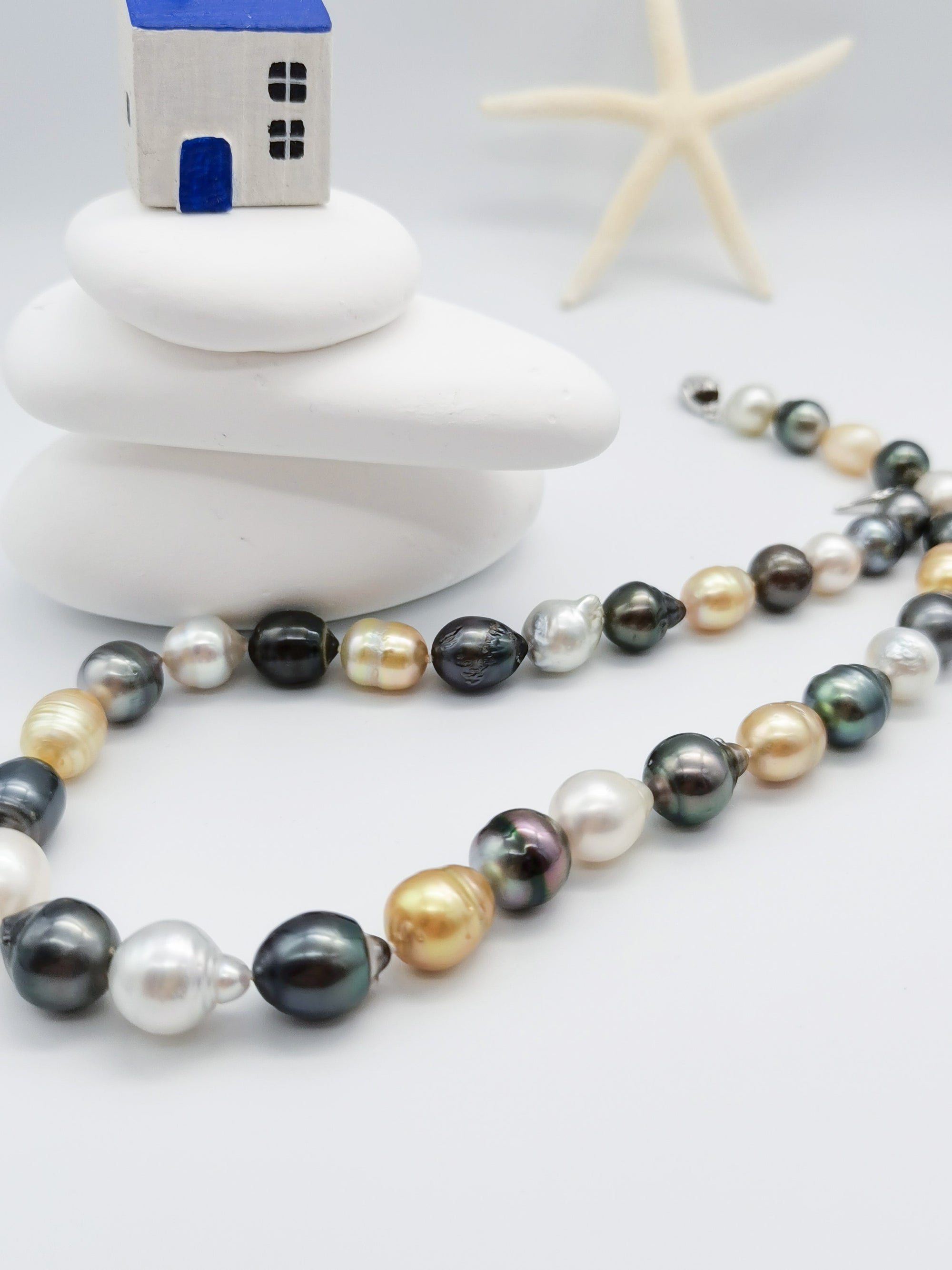 Multicoloured baroque pearl strand necklace artistically arranged with decorative blue house ornament and starfish on stacked white stones in coastal-themed styling