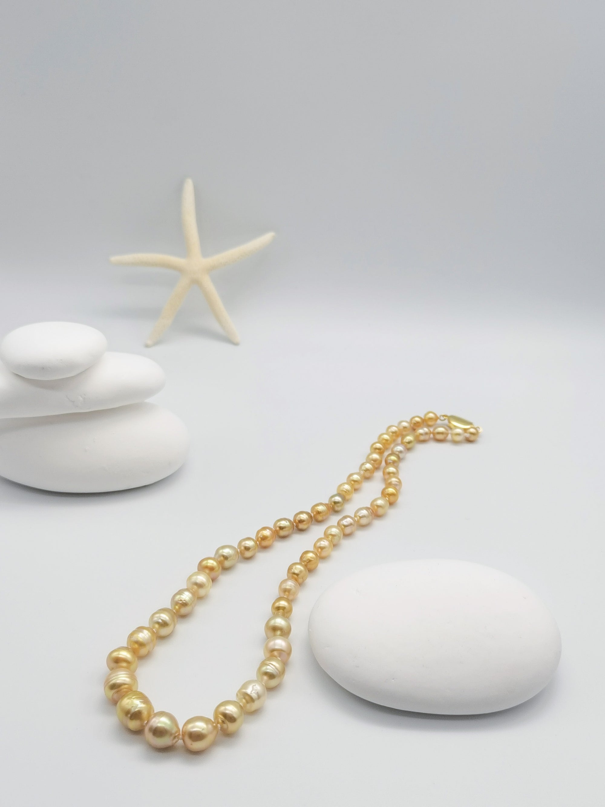 Golden baroque pearl necklace gracefully draped on stacked white stones with starfish in zen-like composition, showcasing the full length and graduated design