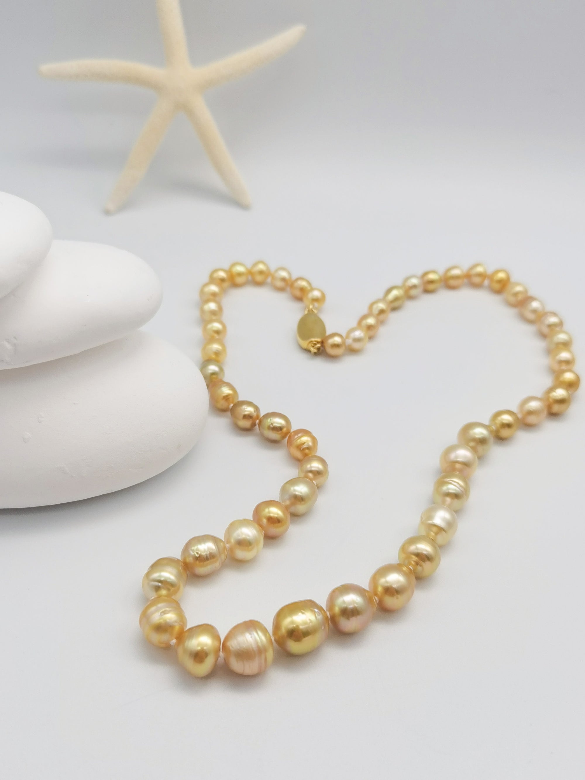 Golden baroque pearl necklace artistically arranged on stacked white stones with starfish backdrop in coastal-themed styling, showcasing the graduated pearl design