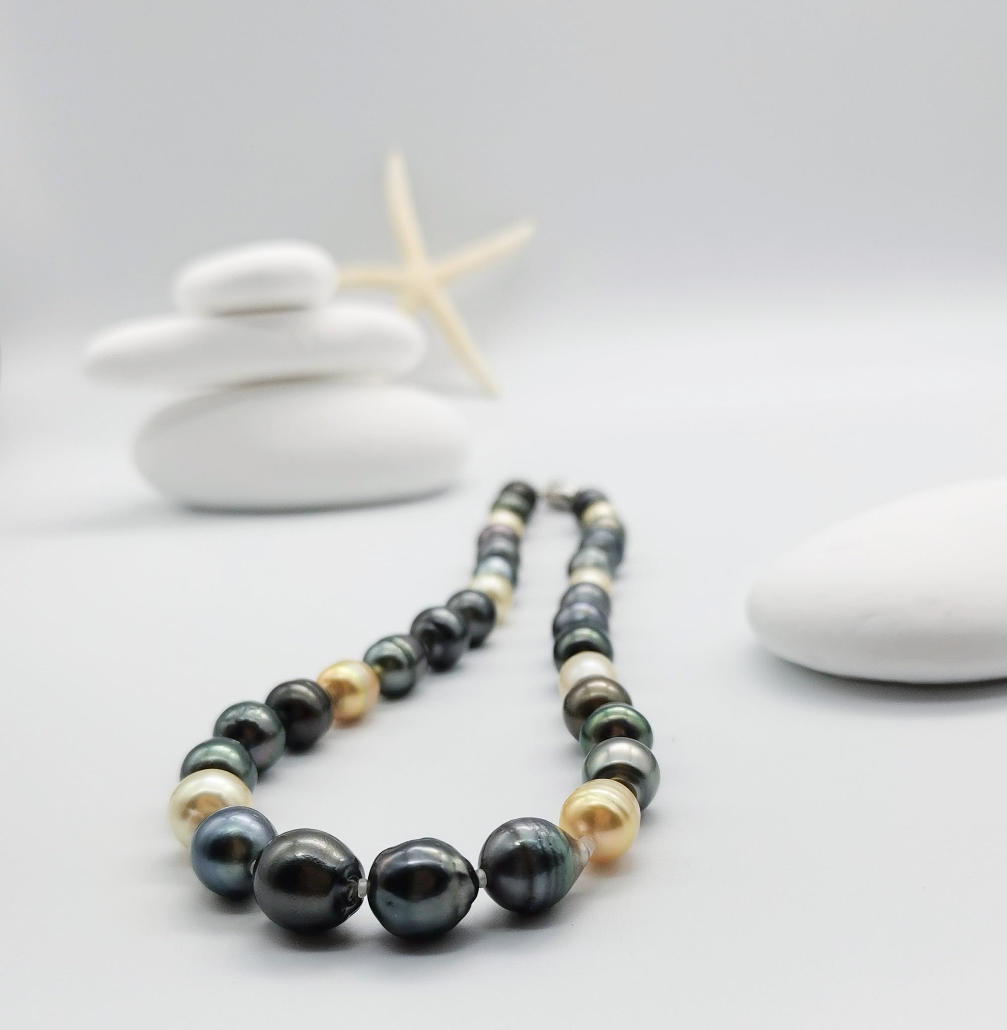 Multicoloured baroque pearl strand necklace artistically arranged in front of stacked white stones with starfish backdrop, displaying the beautiful mix of dark and golden pearl tones