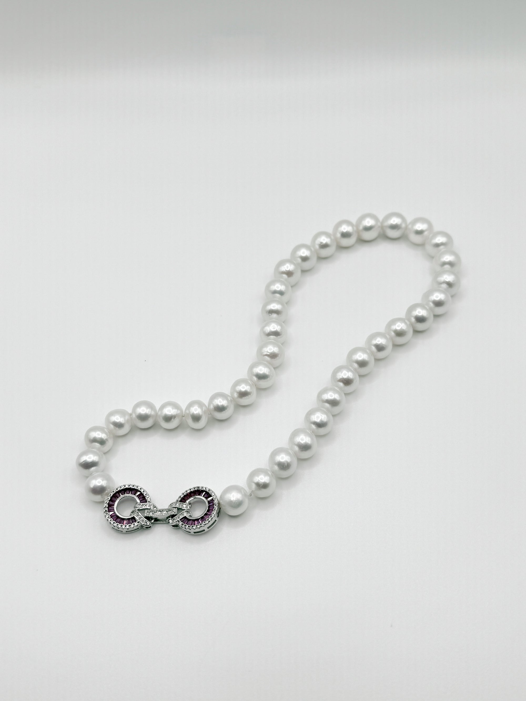 White freshwater pearl necklace with decorative infinity clasp showcasing featuring red and clear CZ setting details and intricate metalwork, elegantly arranged in flowing curve