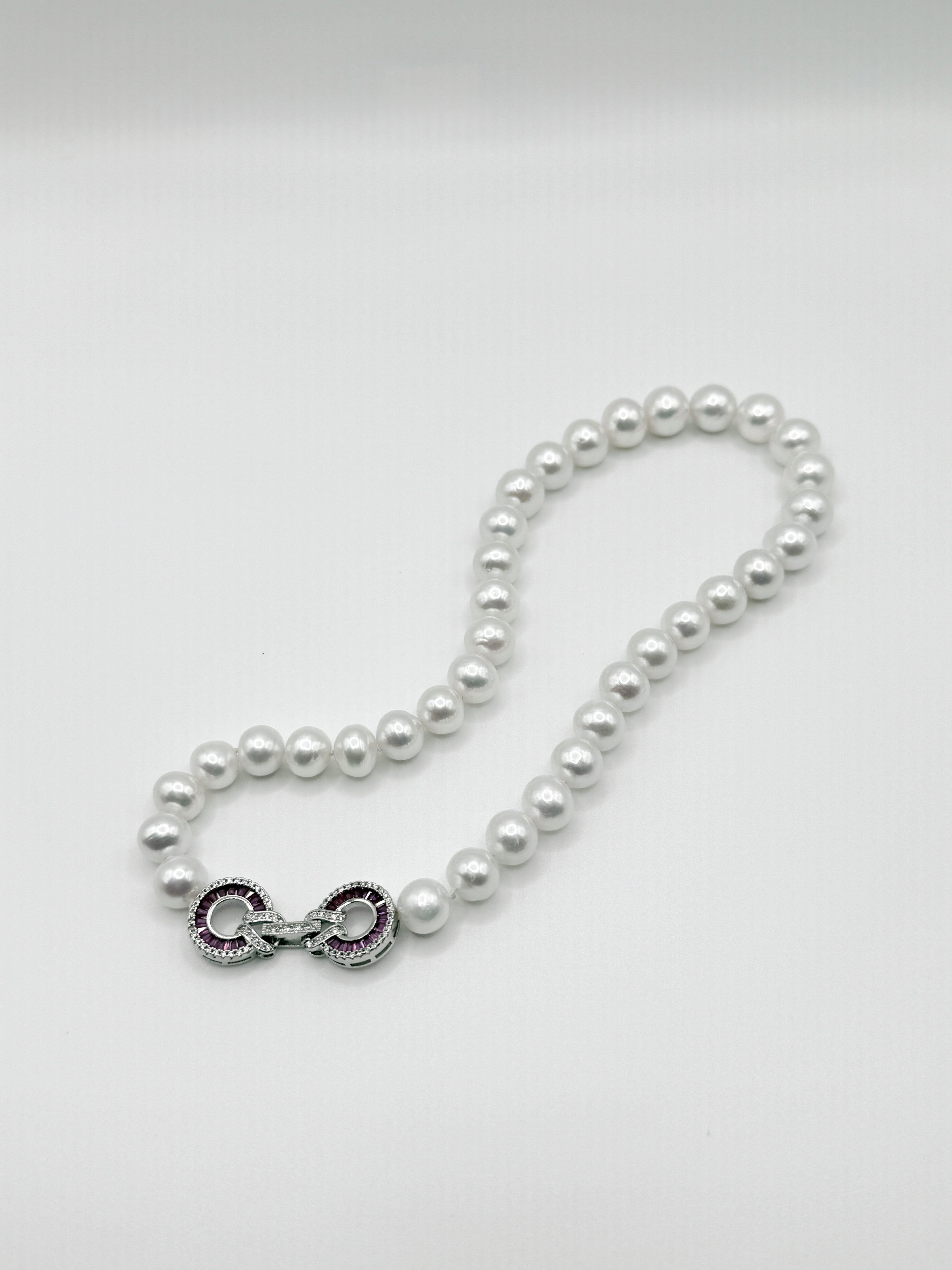 White freshwater pearl necklace with decorative infinity clasp showcasing featuring red and clear CZ setting details and intricate metalwork, elegantly arranged in flowing curve