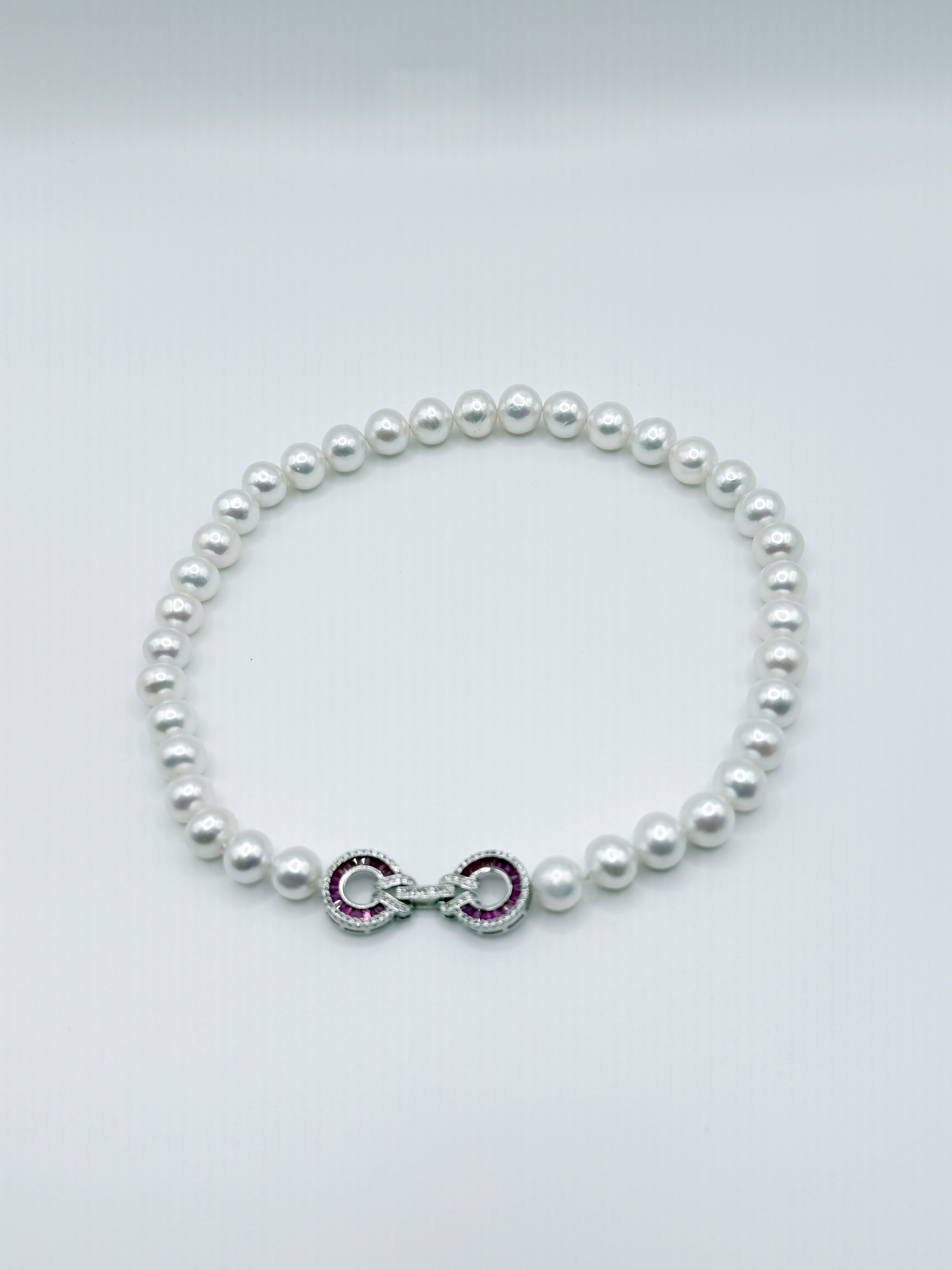 White freshwater pearl strand necklace with ornate infinity clasp featuring red and clear CZ setting details, displayed in circular formation on clean white background