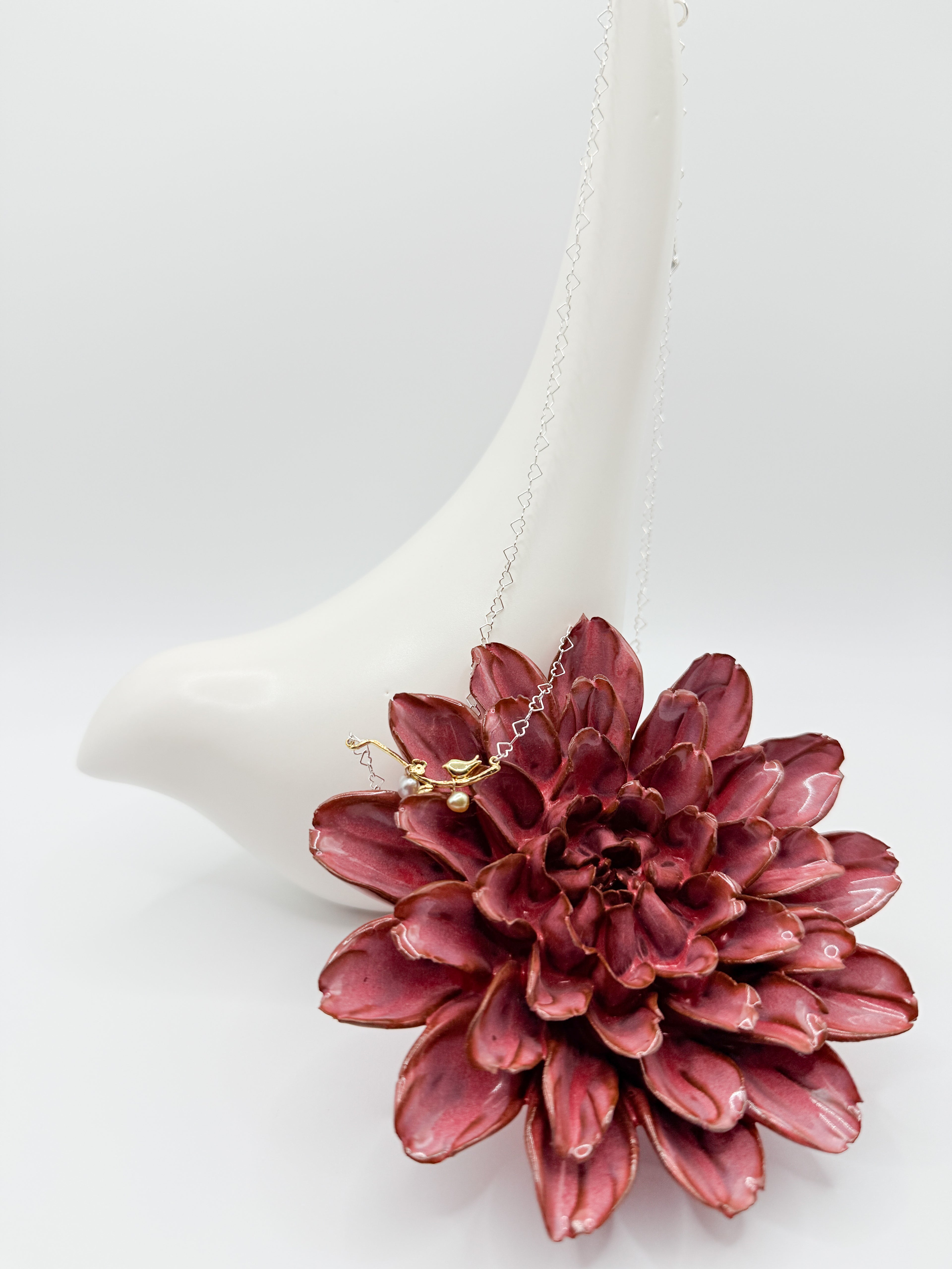 Necklace with bird-on-branch gold pendant and heart-shaped pearls displayed on ceramic bird and flower backdrop