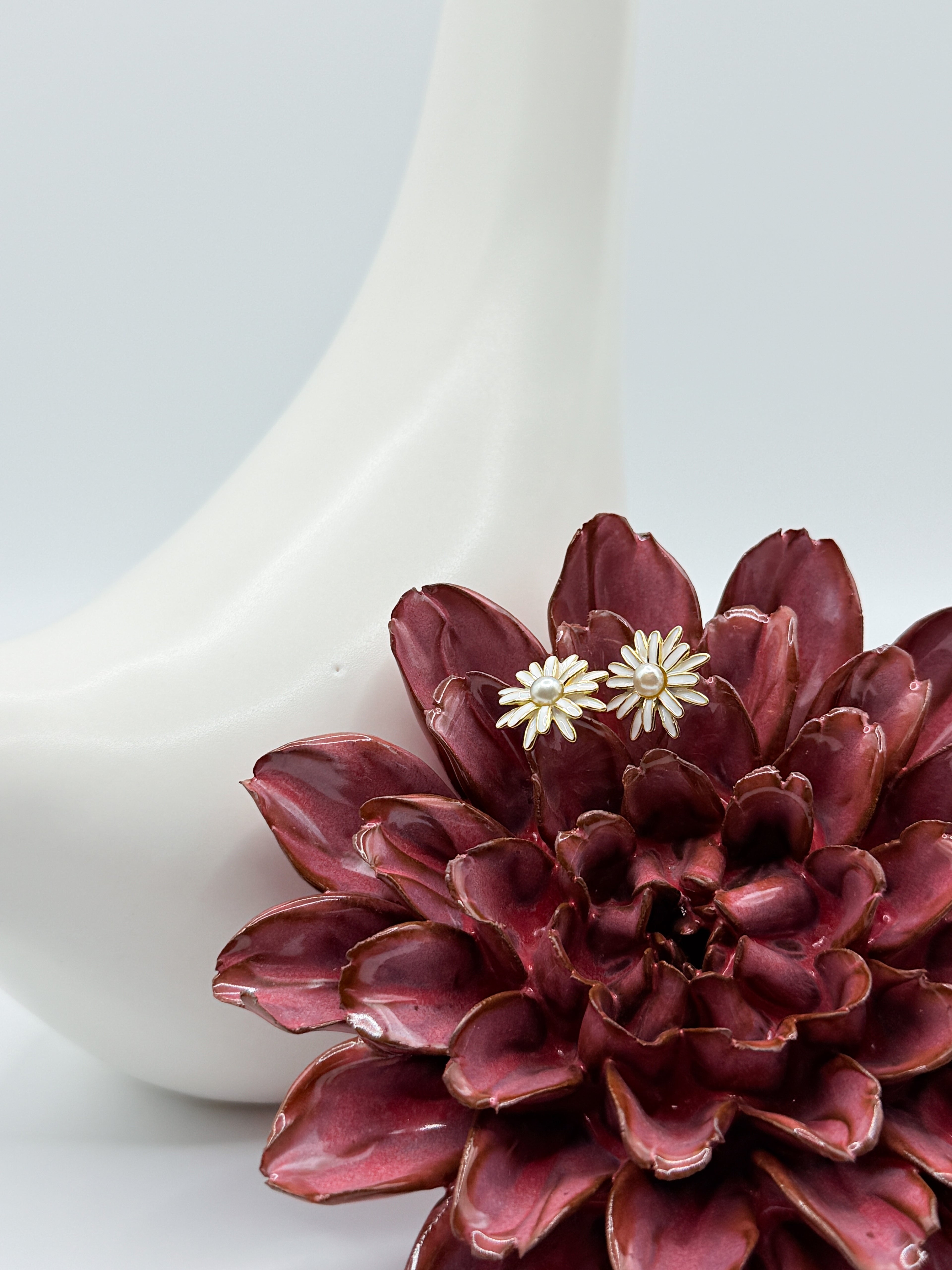 Enamel daisy stud earrings with South Sea Keshi pearls displayed on burgundy ceramic flower and white bird prop