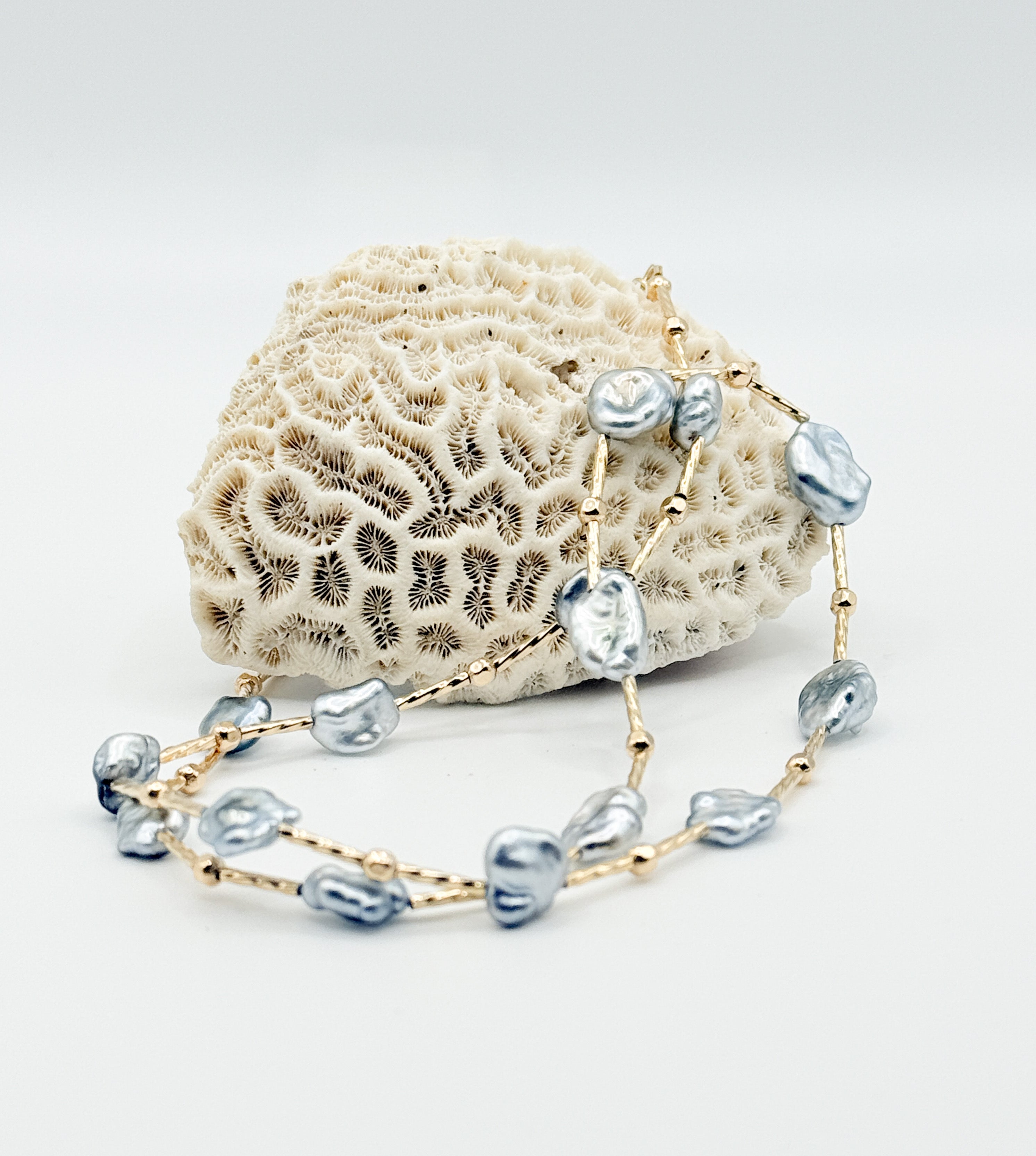 Close up view of blue Tahitian Keshi pearl necklace displayed on white coral, highlighting gold-filled bar and round beads.