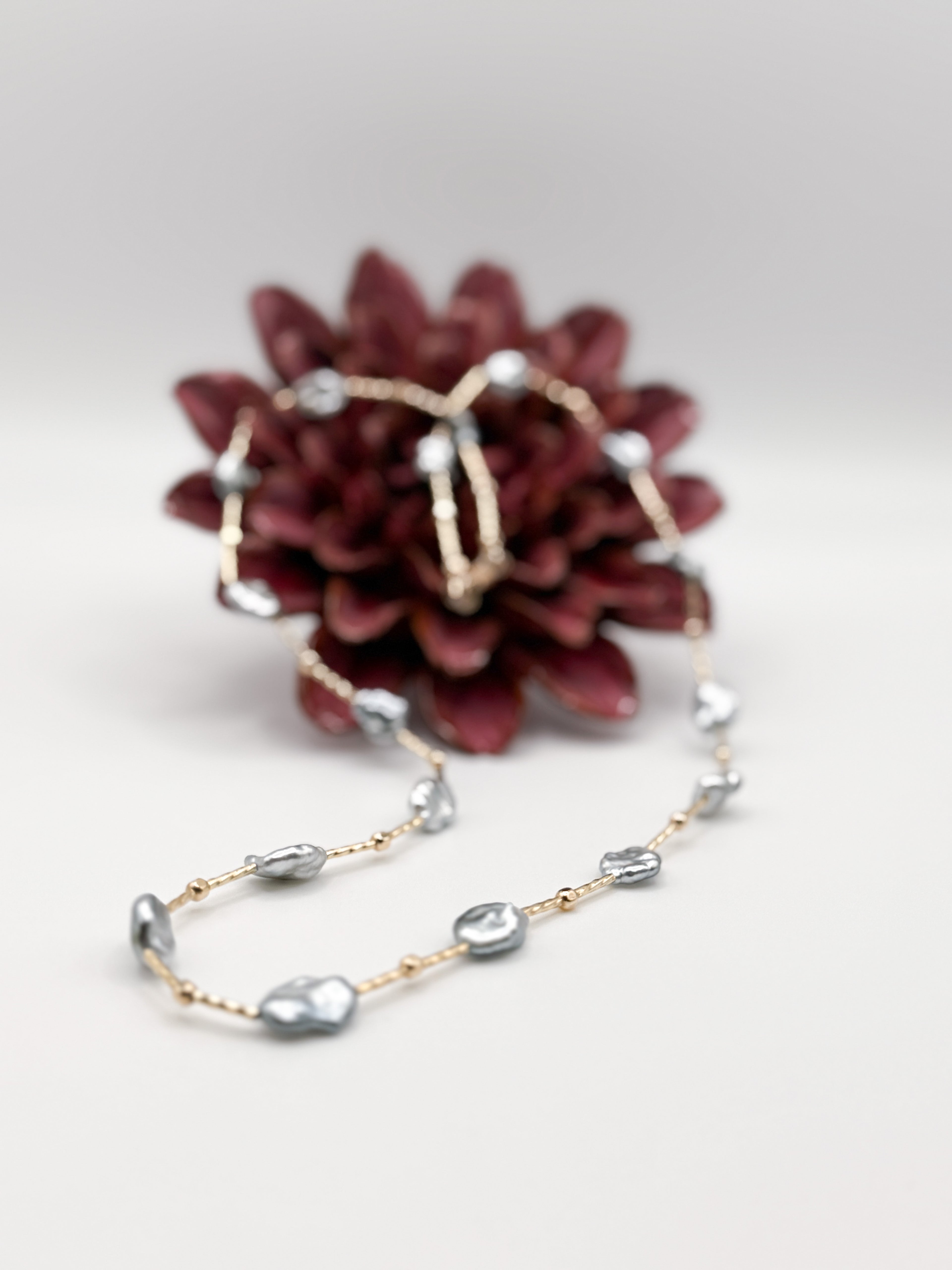 Tahitian Keshi pearl station necklace with gold-filled beads, styled beside red flower for contrast.