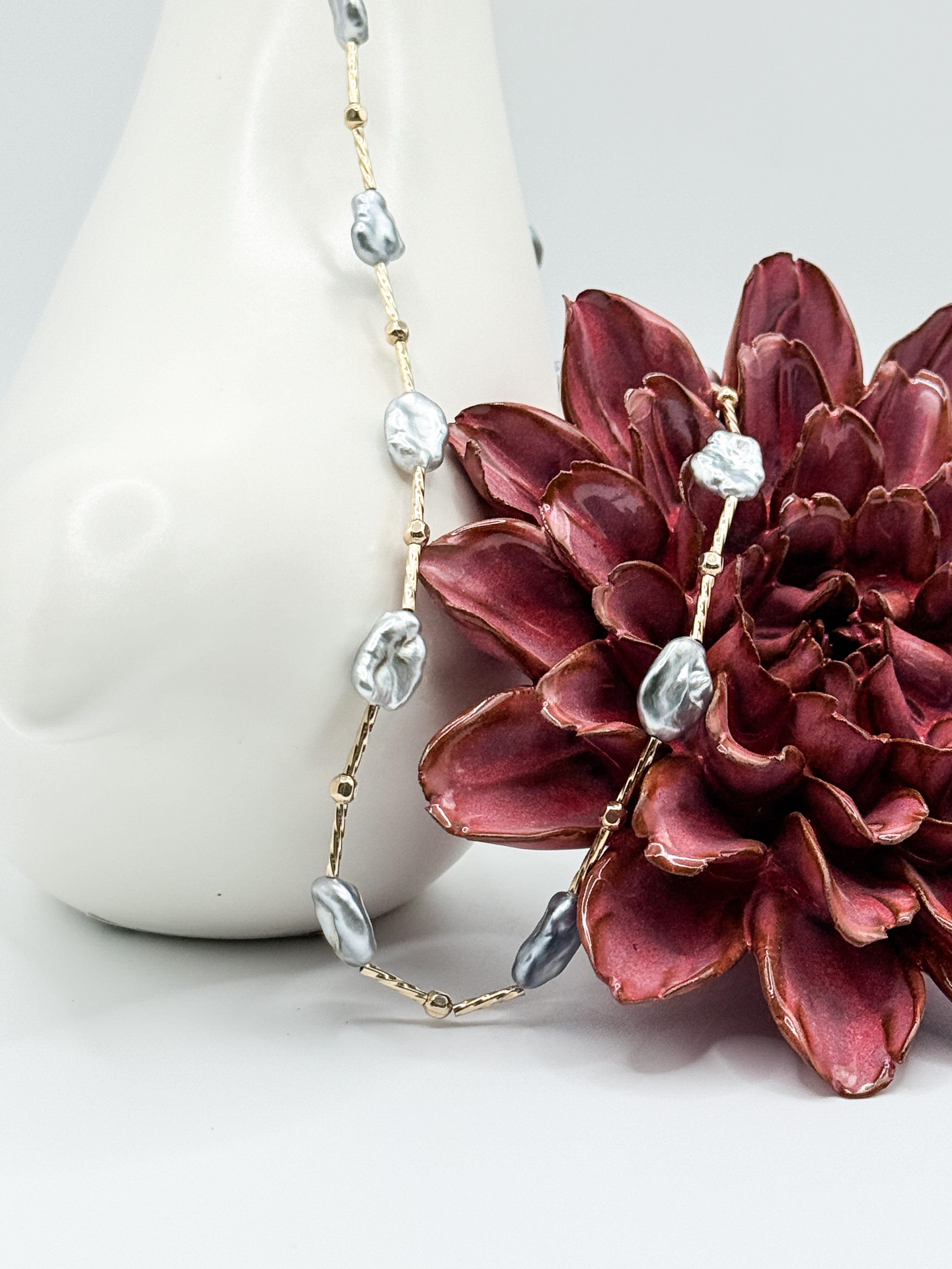 Close-up of blue Tahitian Keshi pearl necklace with gold-filled beads, styled with ceramic vase and red flower