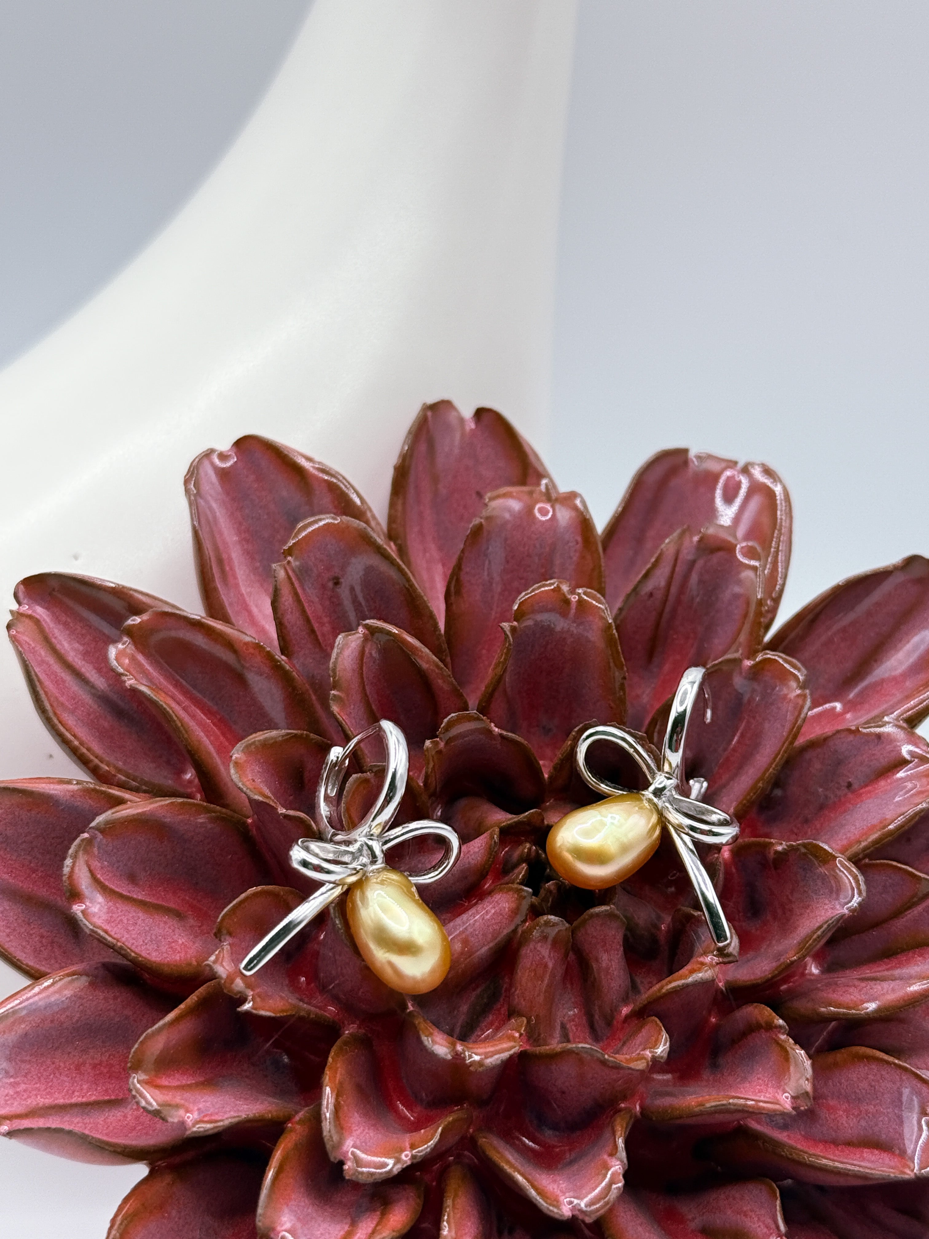 Golden South Sea Keshi pearl bow earrings on red flower, showcasing lustrous pearls and 925 silver looped ribbon setting.