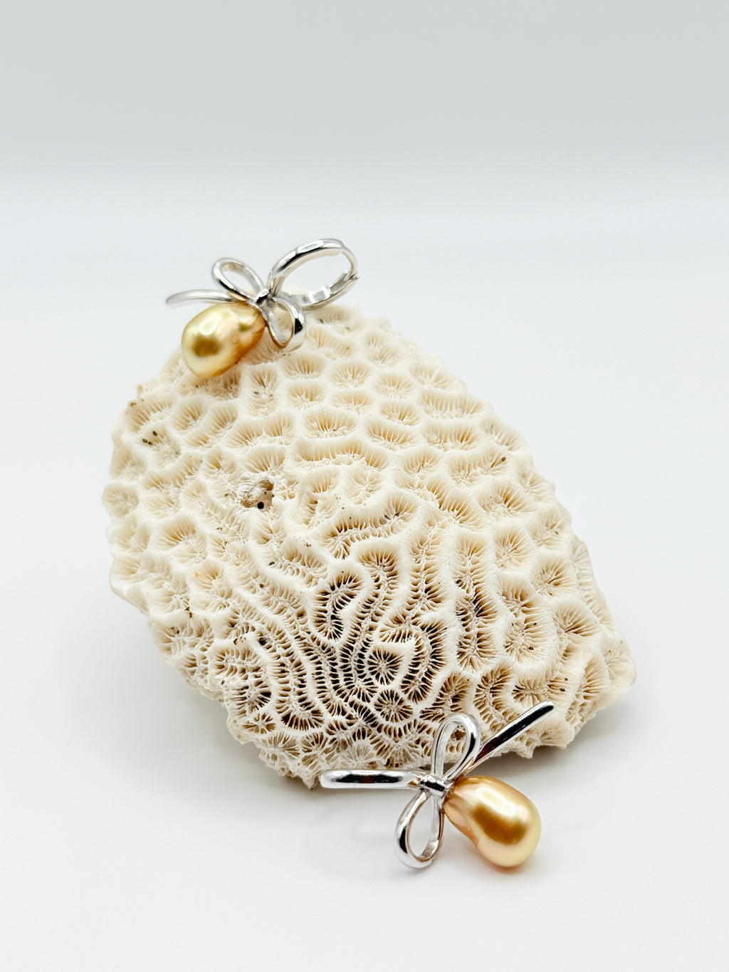 Close-up of golden South Sea Keshi pearl earrings on white coral, highlighting organic pearl shape and silver bow design.