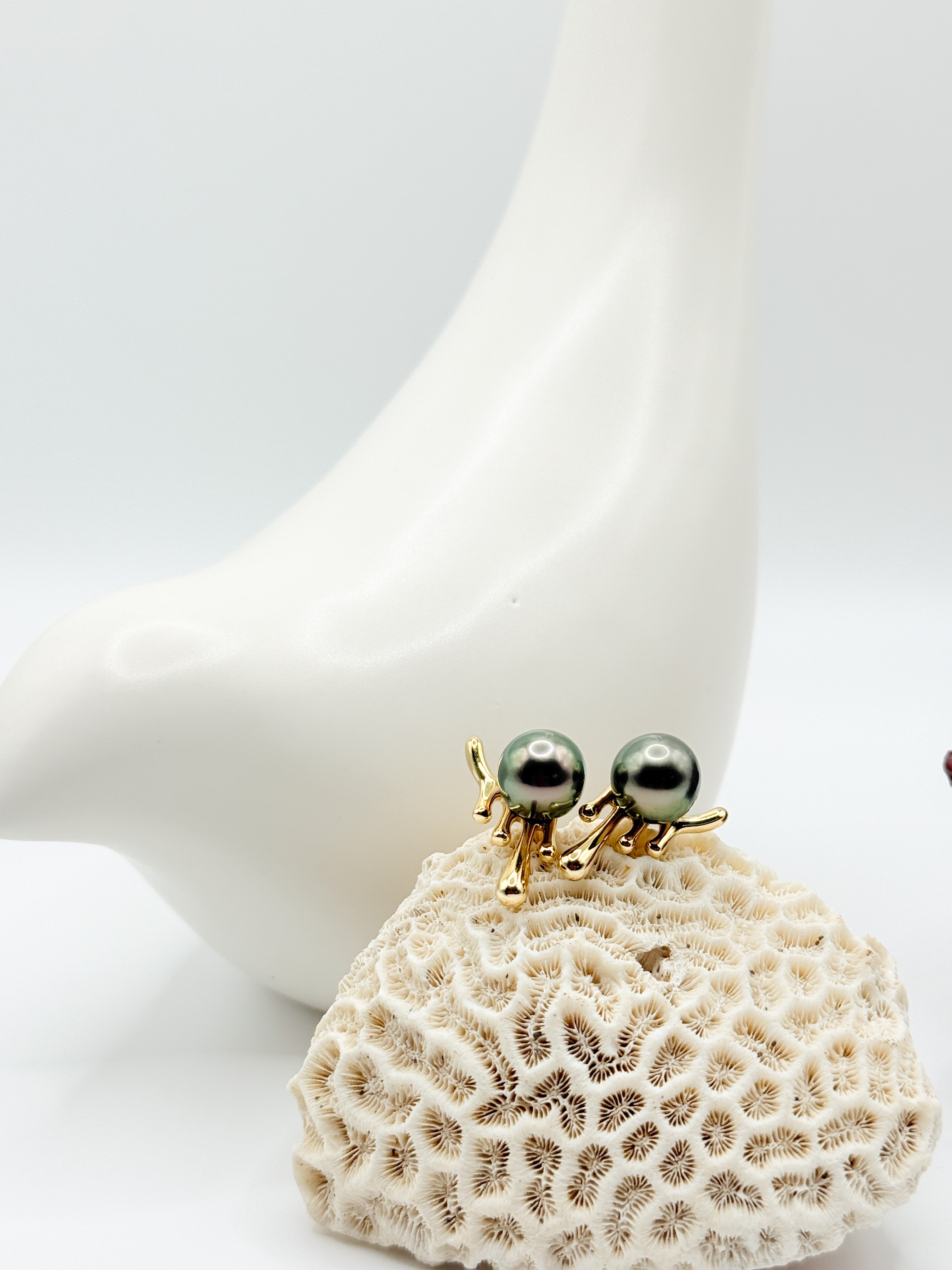 Green Tahitian pearl stud earrings on coral, with molten gold-tone silver setting and ceramic bird background.