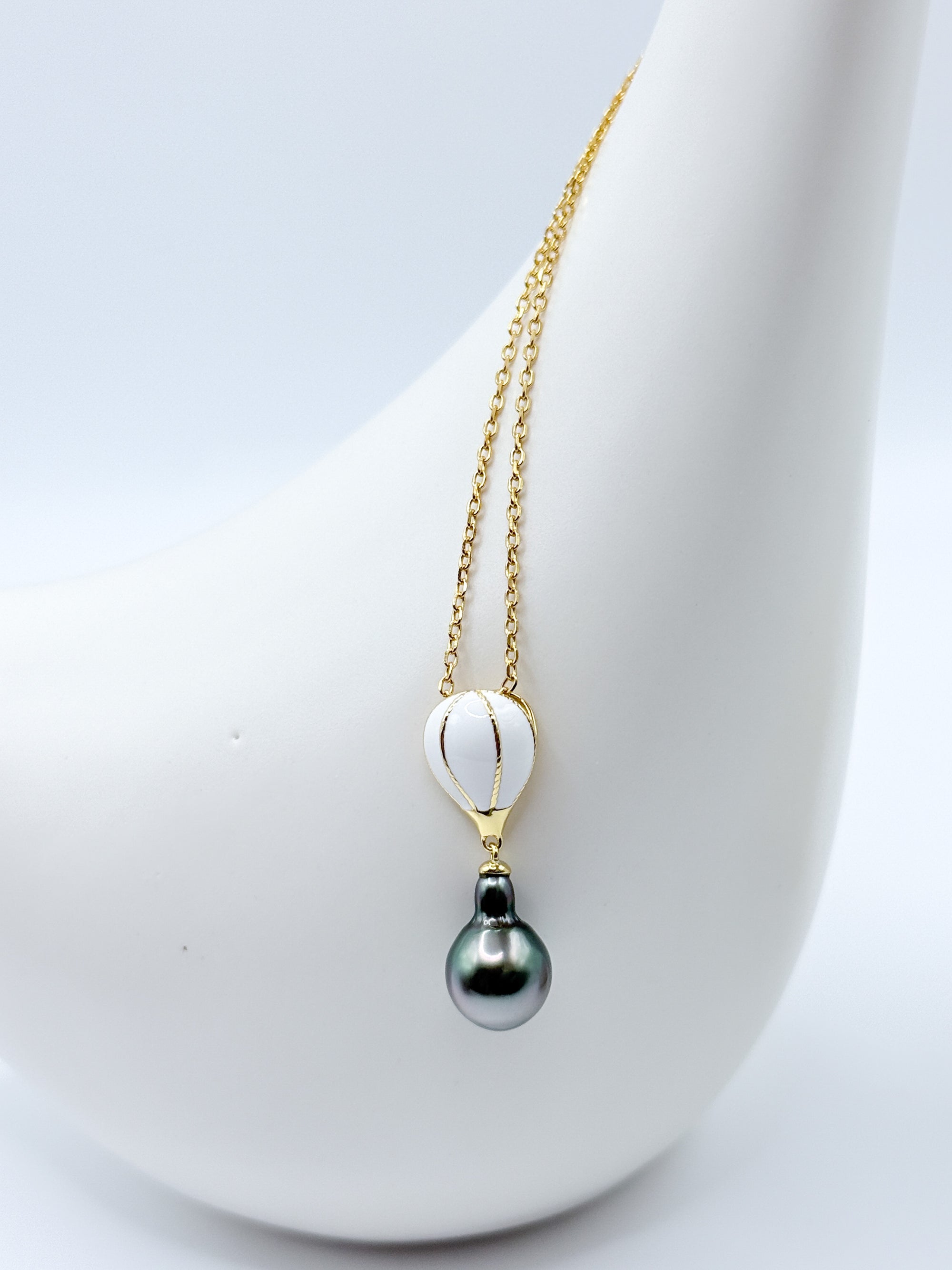 Front-facing shot of Tahitian pearl pendant necklace with hot air balloon charm in white enamel on gold-plated 925 silver, styled on a white ceramic surface.
