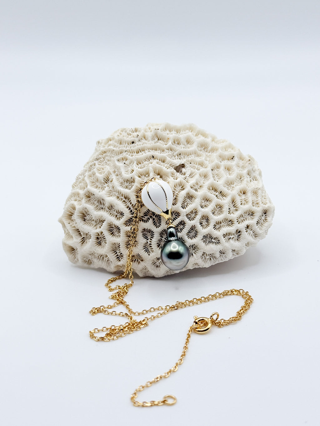 Flat lay of gold chain necklace with whimsical hot air balloon charm and dark Tahitian pearl pendant, arranged on natural coral background.