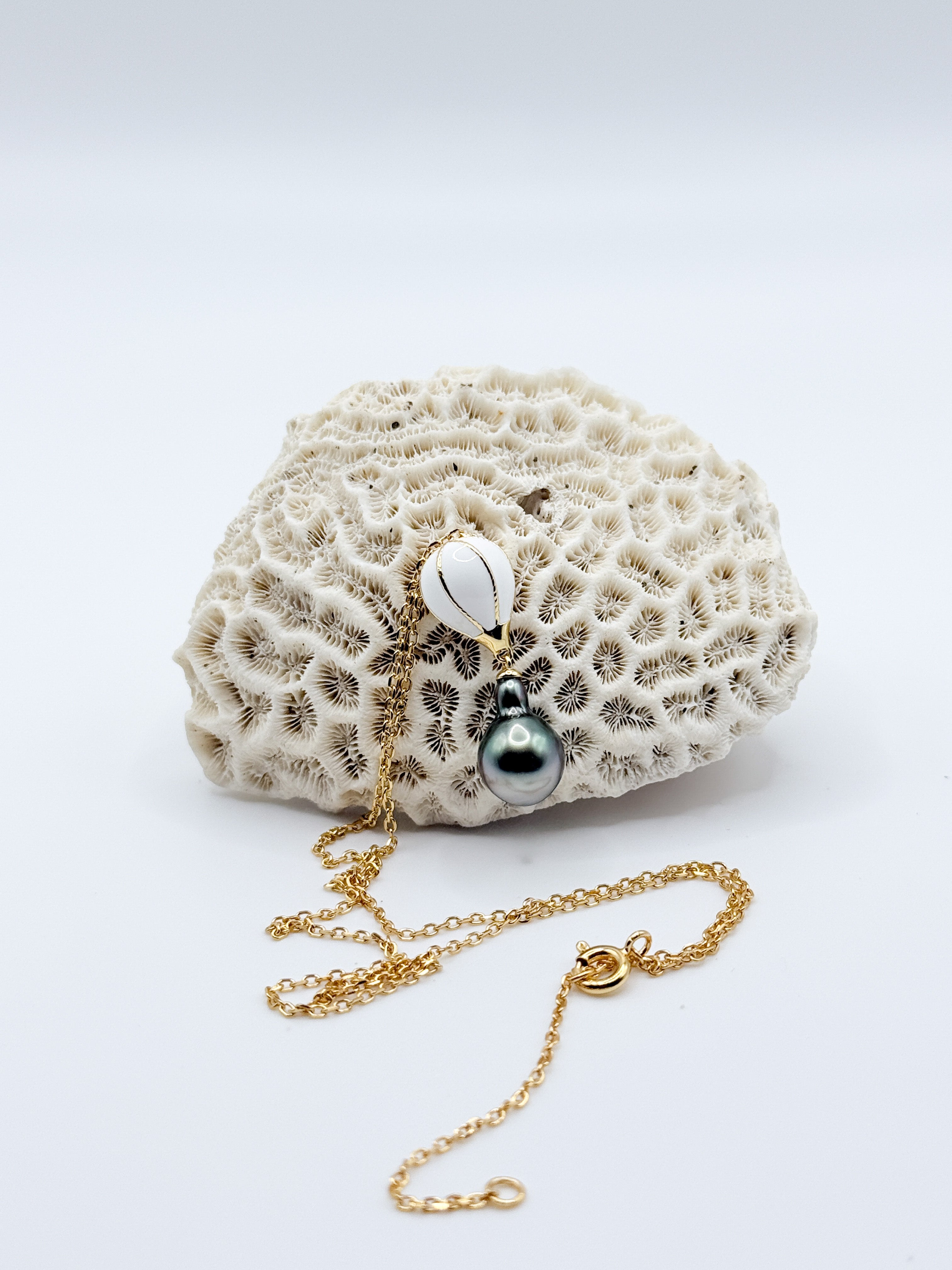 Flat lay of gold chain necklace with whimsical hot air balloon charm and dark Tahitian pearl pendant, arranged on natural coral background.
