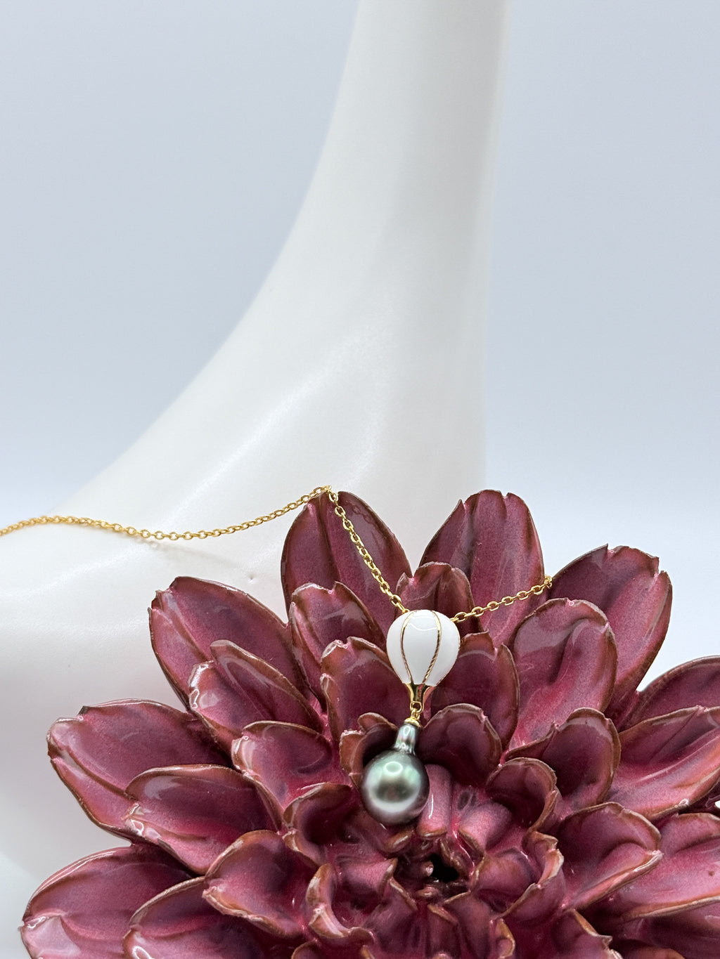 Close-up of gold-plated 925 silver necklace featuring a white enamel hot air balloon charm and drop-shaped Tahitian pearl, displayed on a white ceramic bird and red flower.