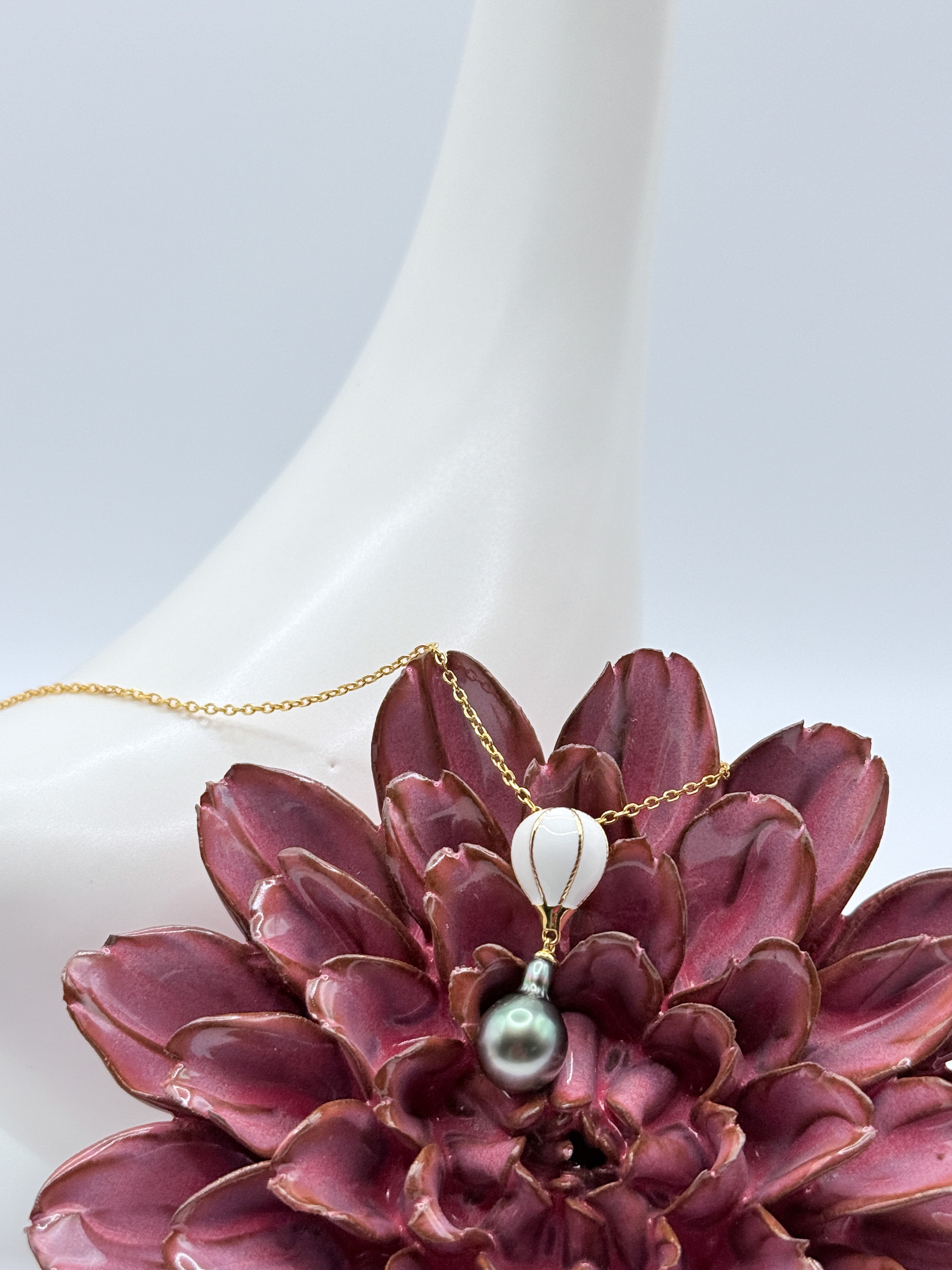 Close-up of gold-plated 925 silver necklace featuring a white enamel hot air balloon charm and drop-shaped Tahitian pearl, displayed on a white ceramic bird and red flower.