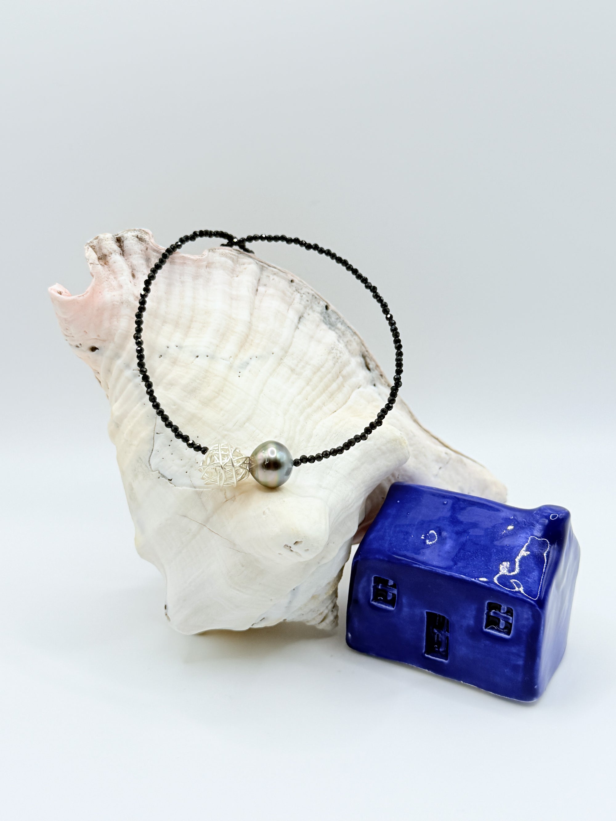 Tahitian pearl choker displayed in circular formation on white conch shell showing full strand of black spinel beads with central baroque pearl and silver wire cage bead