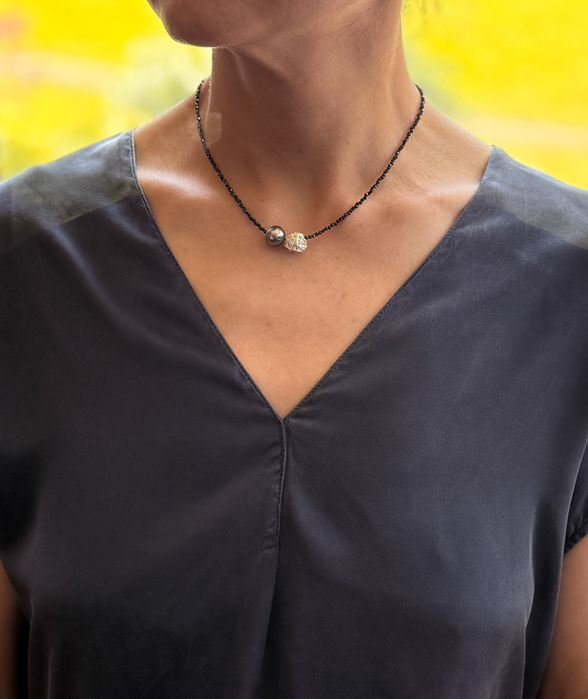 Model wearing Tahitian pearl choker with black spinel beads over charcoal navy blue v-neck top, showing choker length positioning at collarbone with warm yellow background