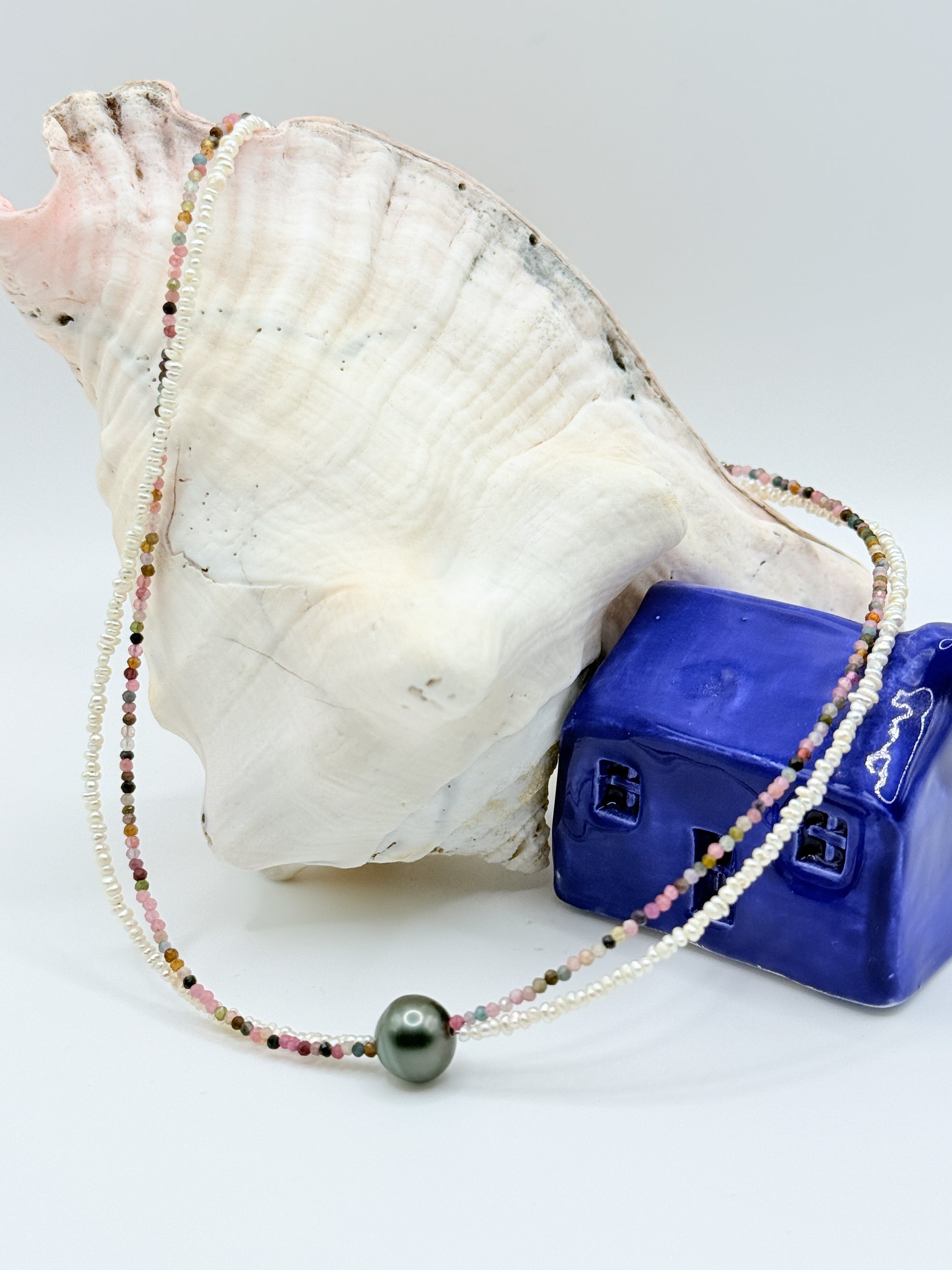 Tahitian pearl choker with tourmaline and seed pearls arranged in flowing curves around large white conch shell and blue ceramic house, showcasing the full length and colourful bead variety on light grey background