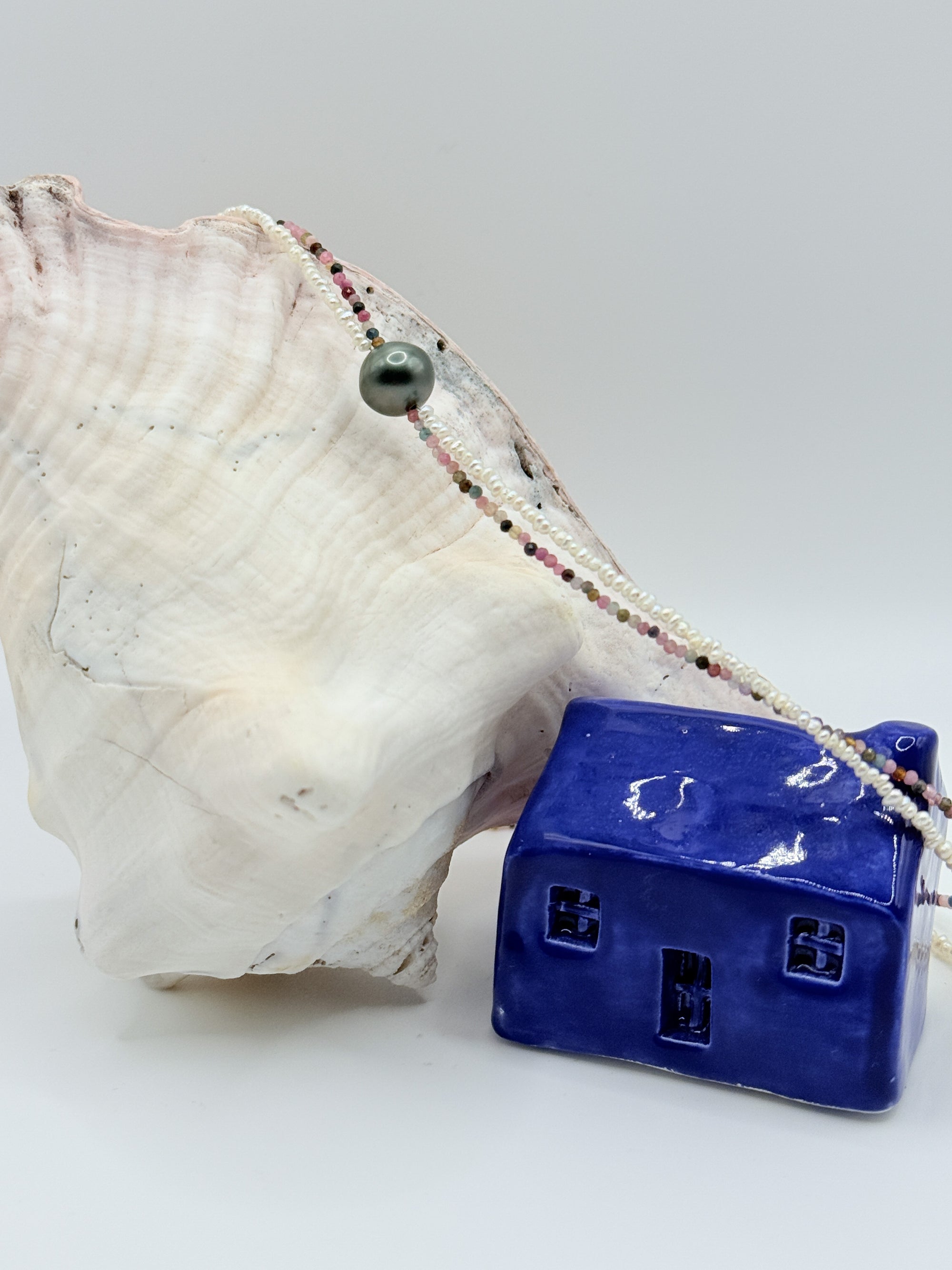 Tahitian pearl choker draped over white conch shell displaying the full strand of colourful tourmaline beads, seed pearls, and central green Tahitian baroque pearl next to blue ceramic house decoration