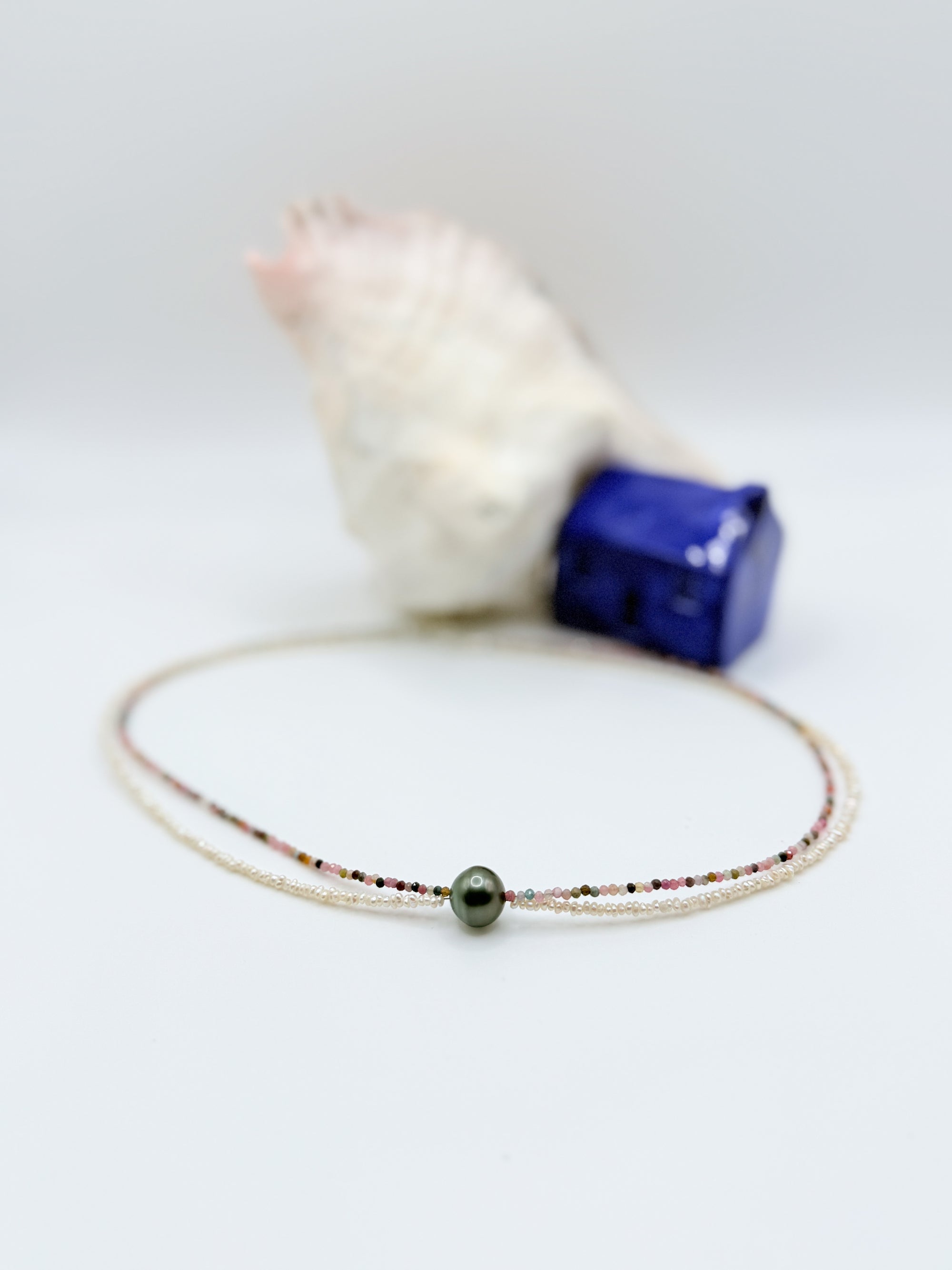 Tahitian pearl choker with tourmaline and seed pearls laid flat in circular formation showing green baroque pearl centrepiece surrounded by multi-coloured faceted tourmaline beads and tiny seed pearls with white shell and blue ceramic house in soft focus background