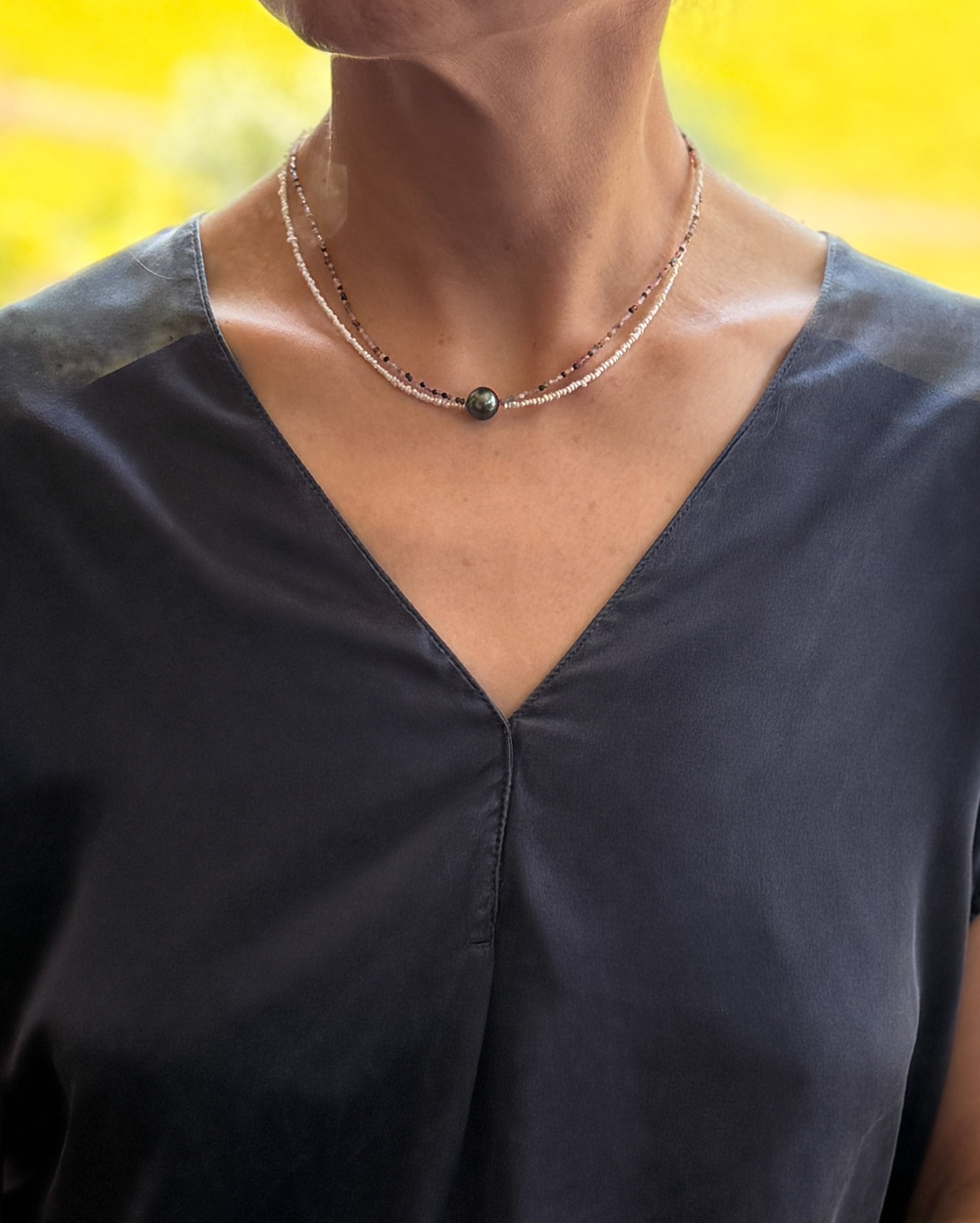 Model wearing Tahitian pearl choker with colourful tourmaline and seed pearl beads over navy-blue v-neck top, showing choker length positioning at collarbone with warm yellow background