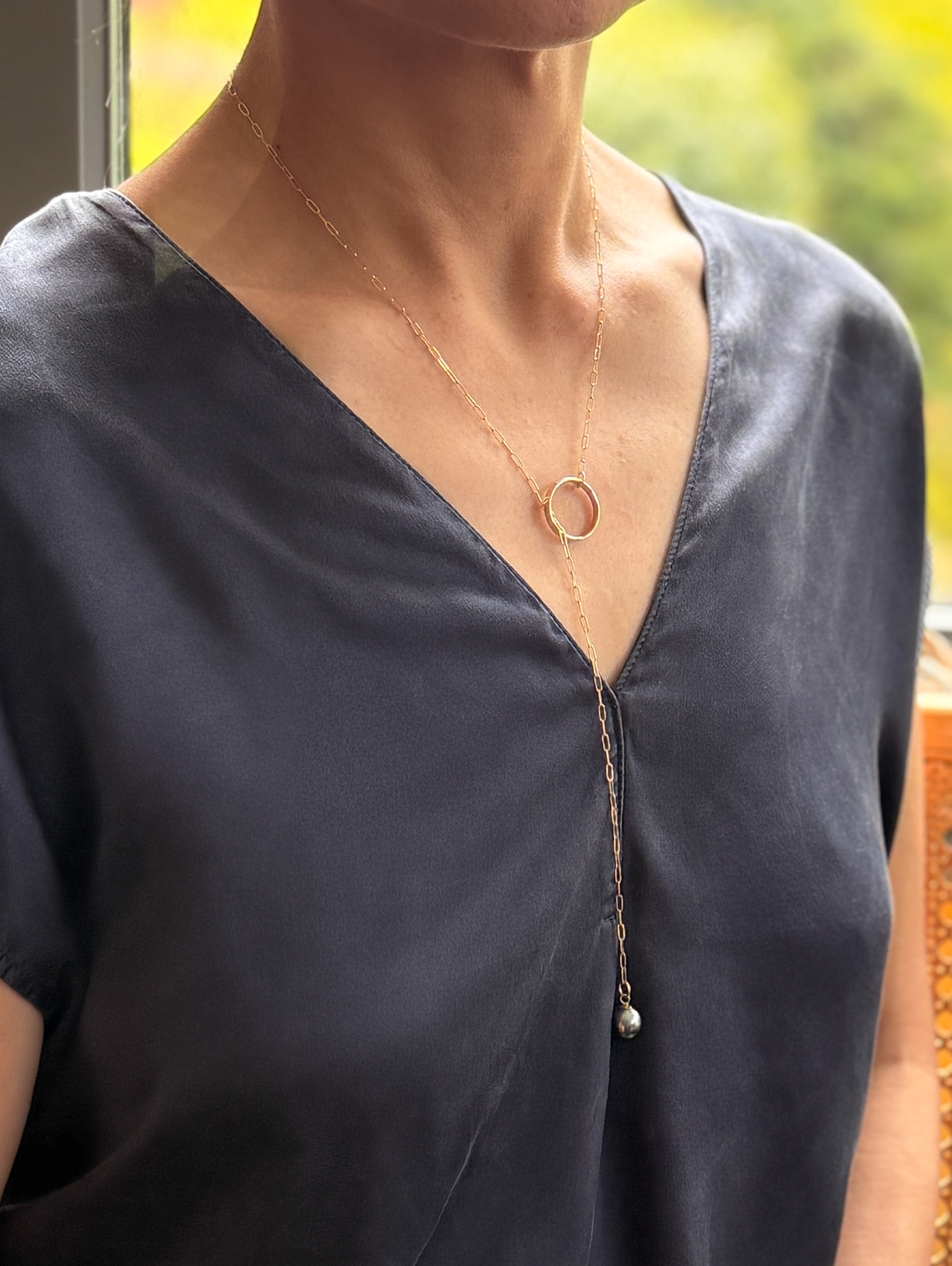 Model wearing gold chain lariat necklace with Tahitian pearl over navy-blue grey v-neck top, demonstrating the adjustable lariat styling with pearl pendant hanging at chest level and warm natural lighting