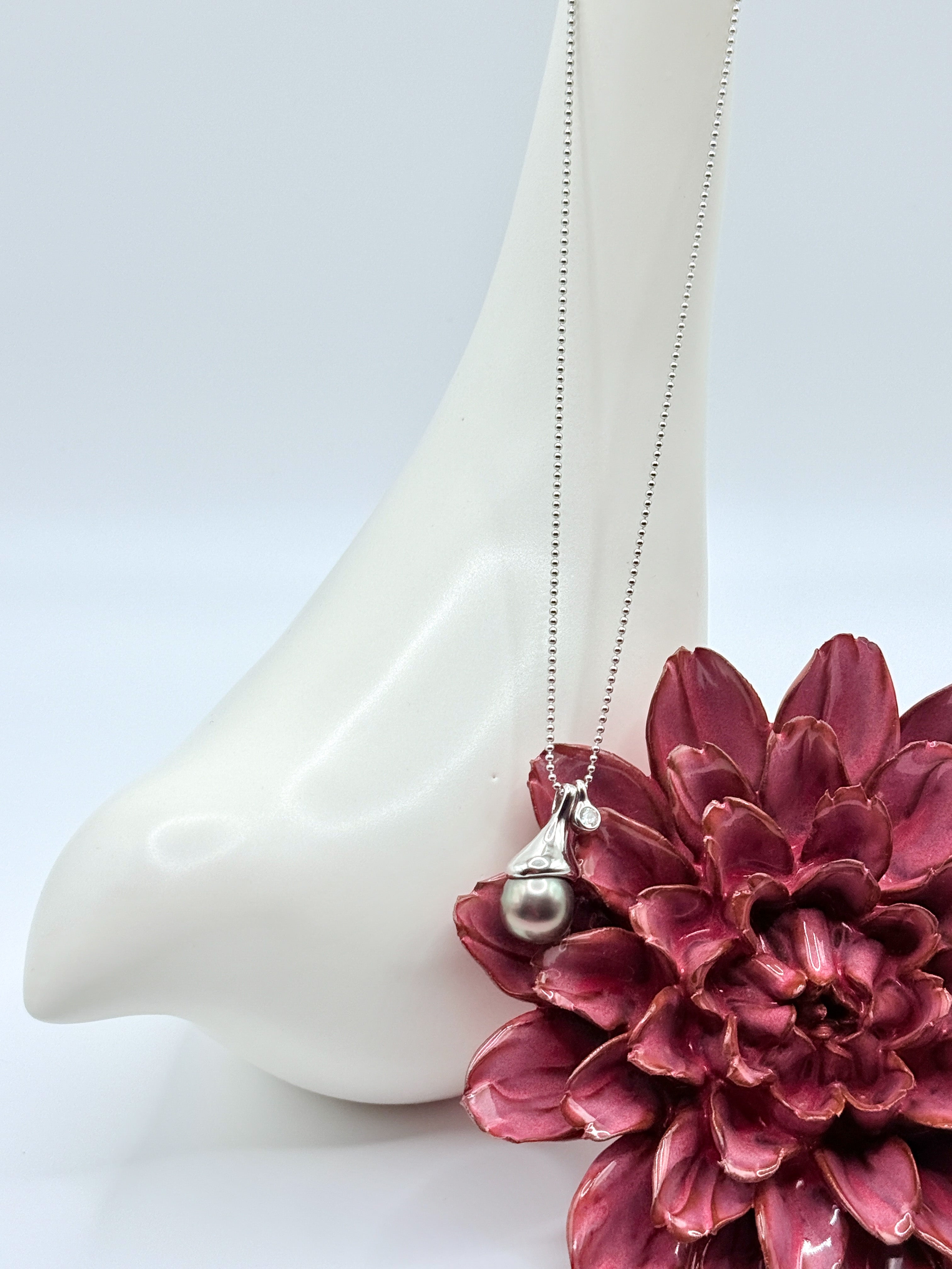 Close-up of sterling silver ball chain necklace showing Tahitian pearl in contemporary silver pendant setting with white ceramic backdrop and burgundy flower