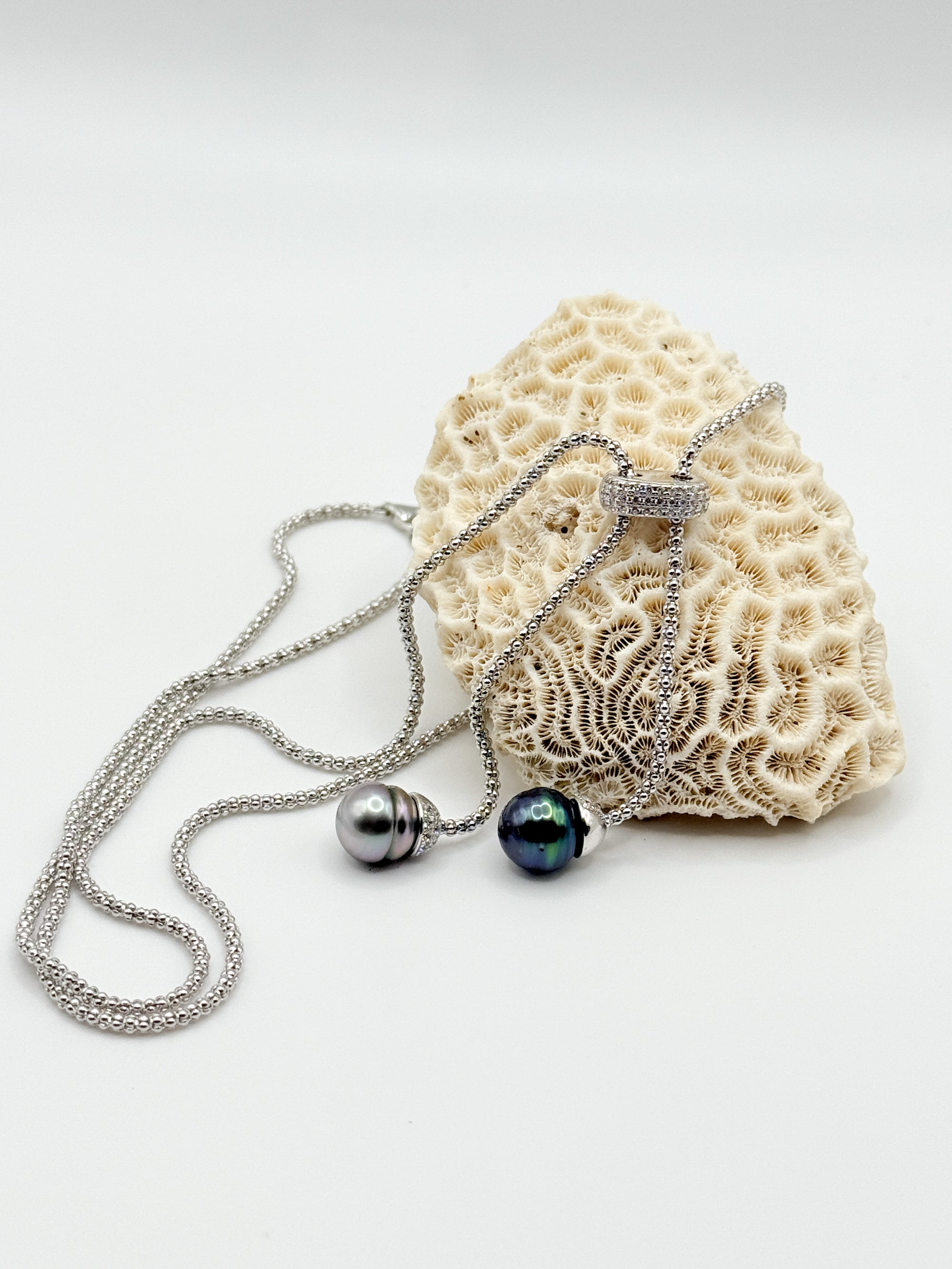 Sterling silver lariat necklace with two Tahitian pearls in different colours displayed on white coral specimen, showing one grey pearl and one dark green pearl with silver accents