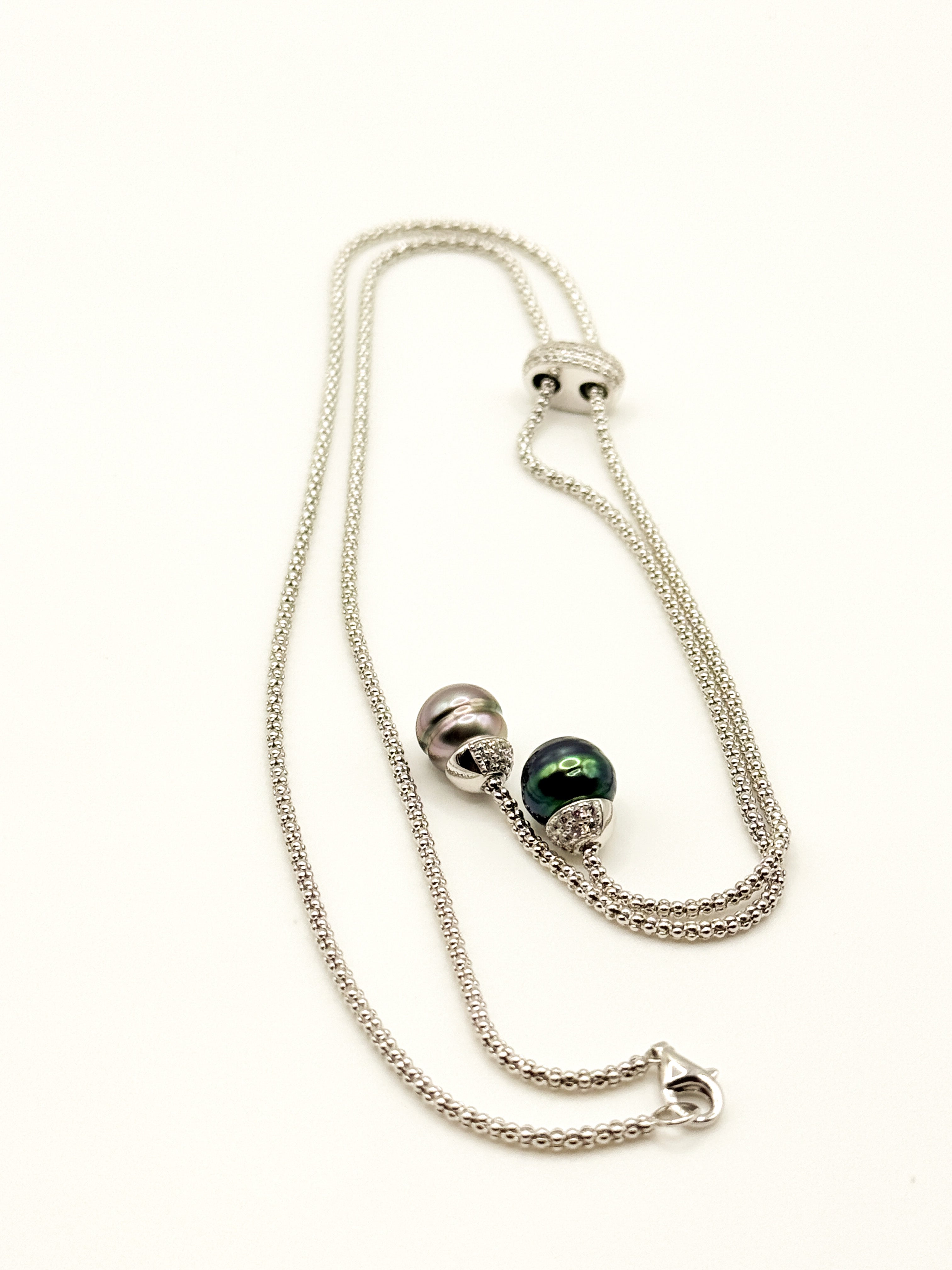 Sterling silver lariat necklace with two Tahitian pearls in different colours displayed on white coral specimen, showing one light pink/aubergine pearl and one vivid green pearl with iridescence