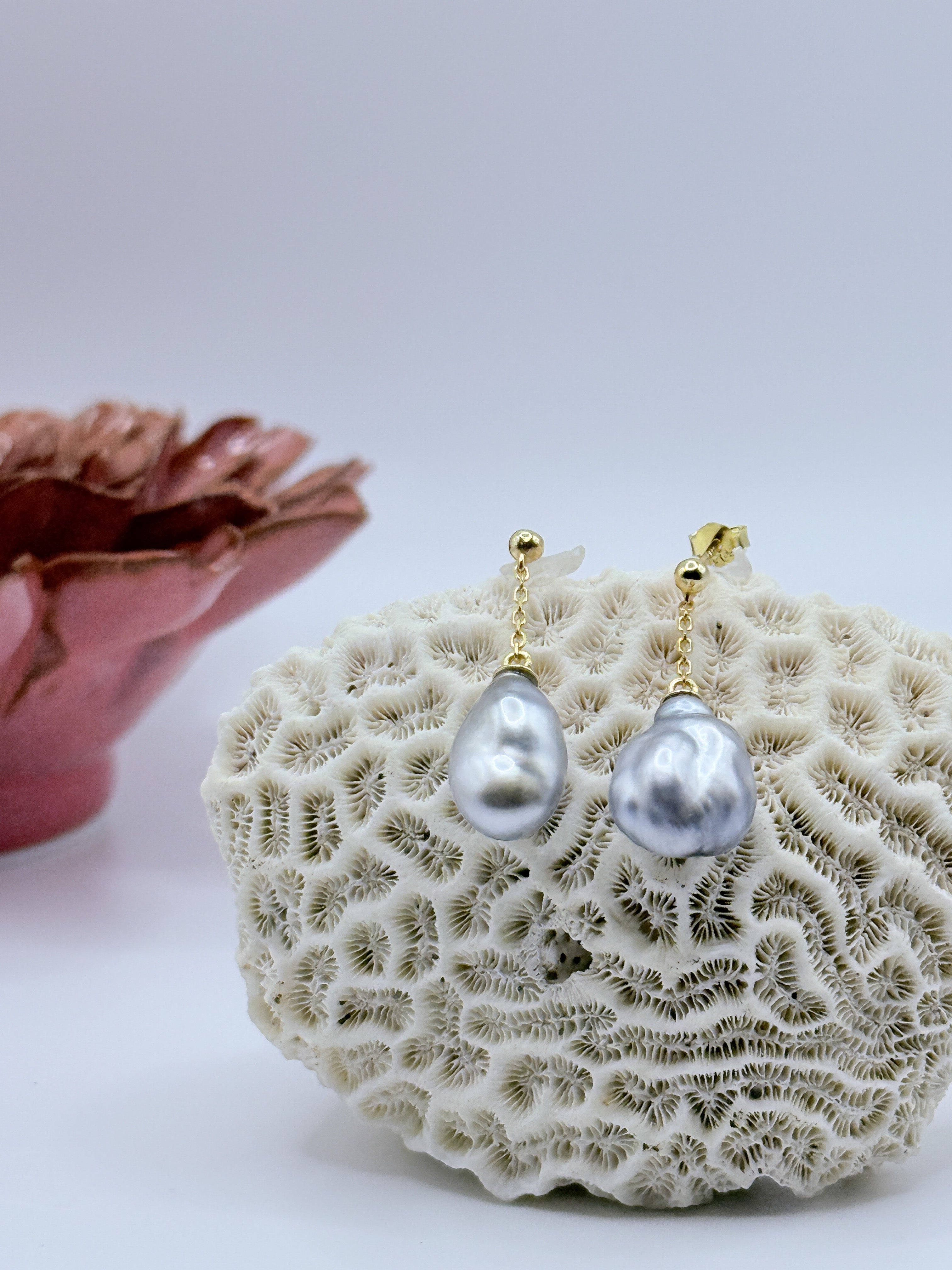 Gold drop earrings with Tahitian keshi pearls displayed on white coral specimen, showing the natural baroque shape and lustrous surface of the pearls with burgundy flower in soft focus background