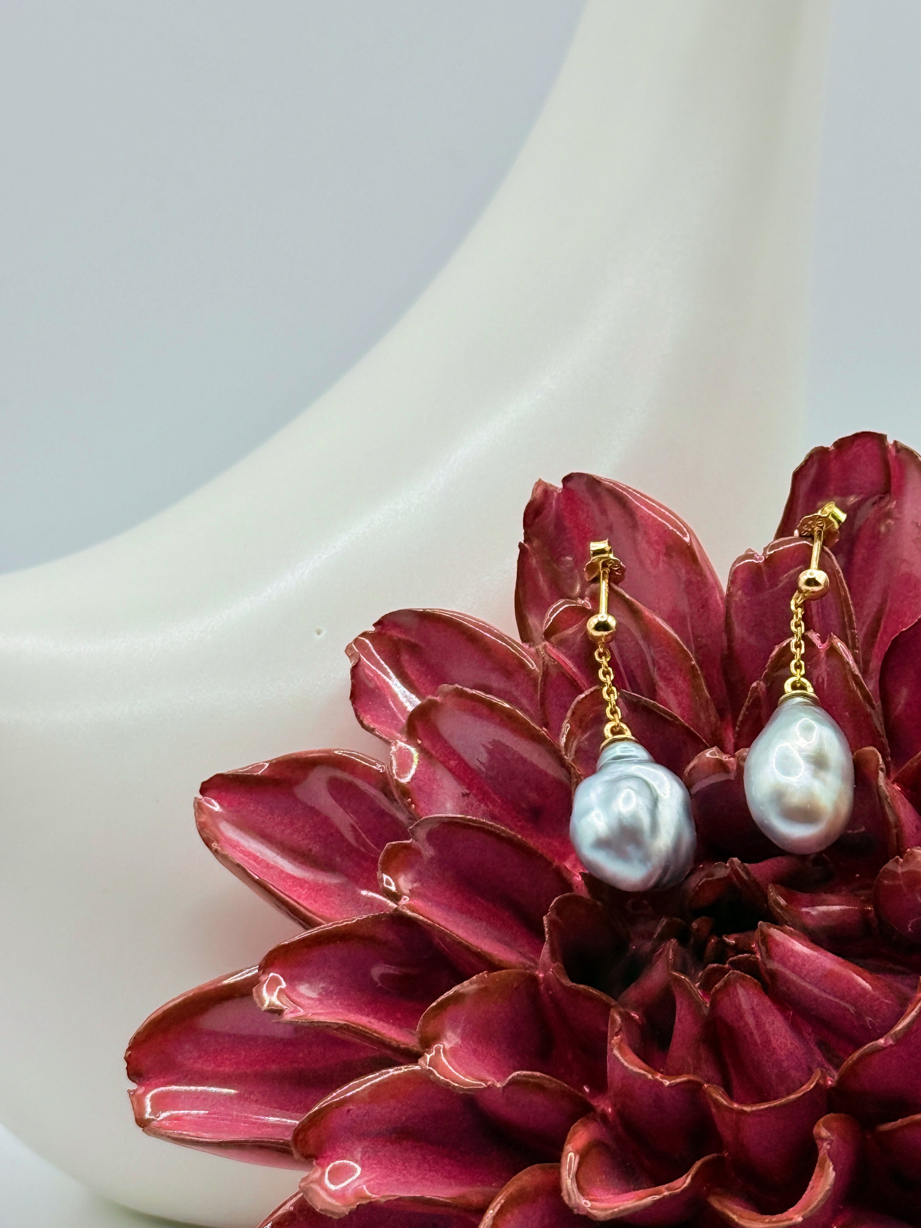 Close-up of gold drop earrings featuring baroque Tahitian keshi pearls hanging from fine gold chains, displayed with burgundy flower against white ceramic backdrop