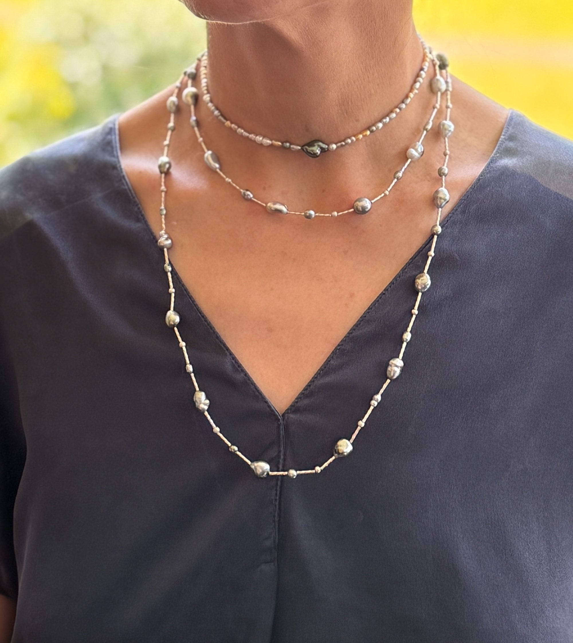Model wearing long sterling silver station necklace with Tahitian keshi pearls in multiple layers over navy-blue v-neck top, showing versatile styling with warm natural lighting