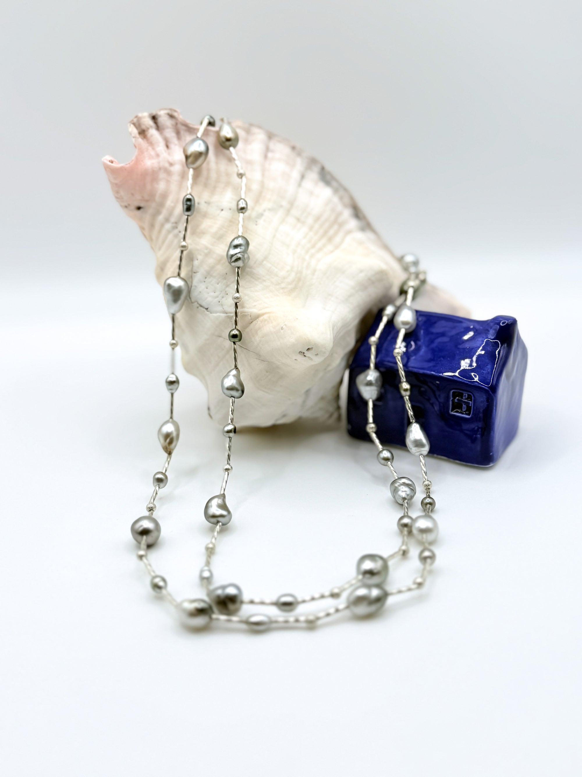 Long sterling silver station necklace with mixed Tahitian keshi pearls draped over white conch shell and blue ceramic house, displaying the full length and variety of pearl colours and shapes
