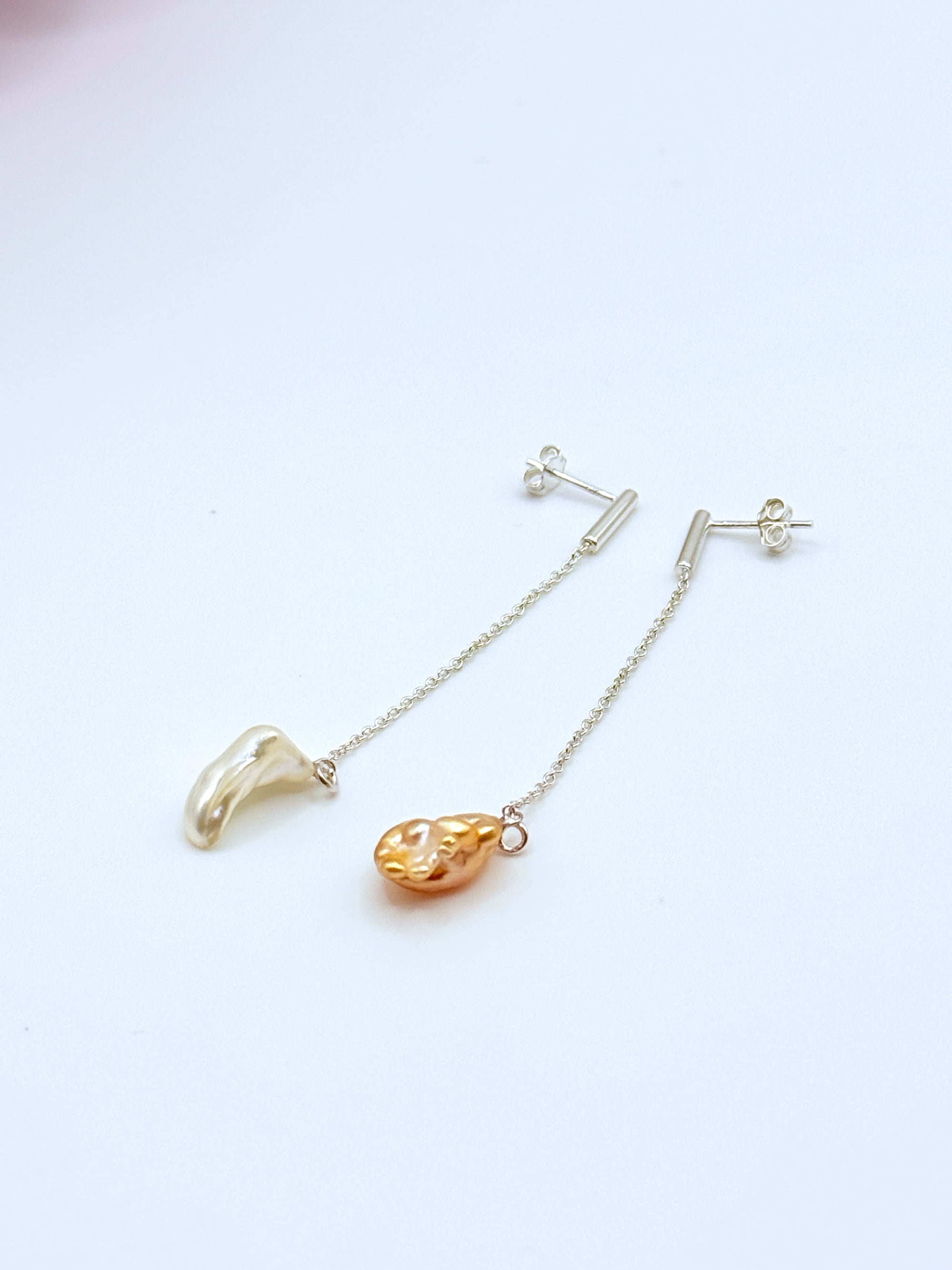 Close-up detail of asymmetrical gold drop earrings with South Sea keshi pearls on white background, showing the delicate gold chain construction and bar-style ear posts