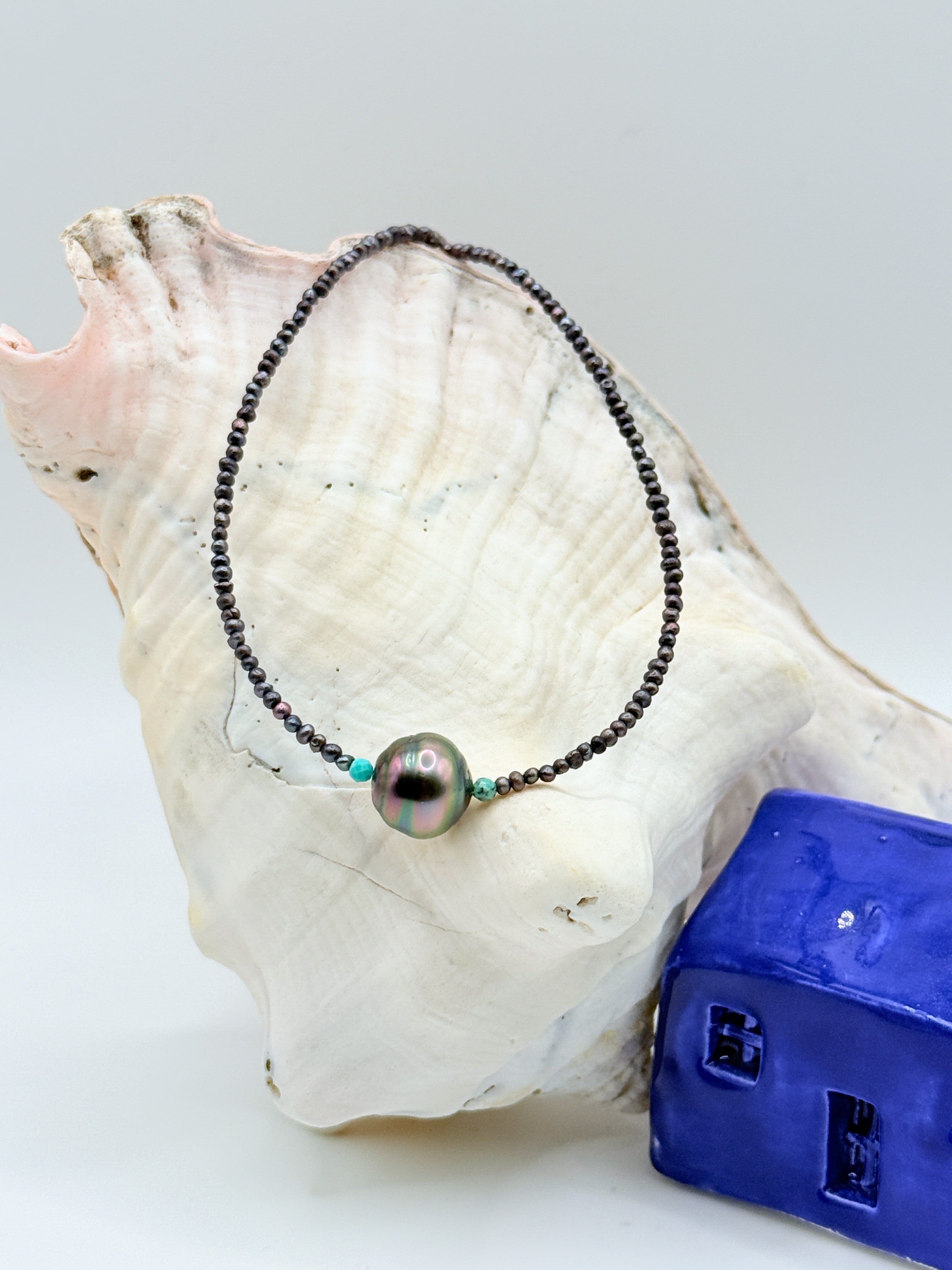 Close-up of delicate choker with dark Tahitian baroque pearl centrepiece flanked by small turquoise accent beads and tiny black seed pearls, displayed on white conch shell with blue ceramic house