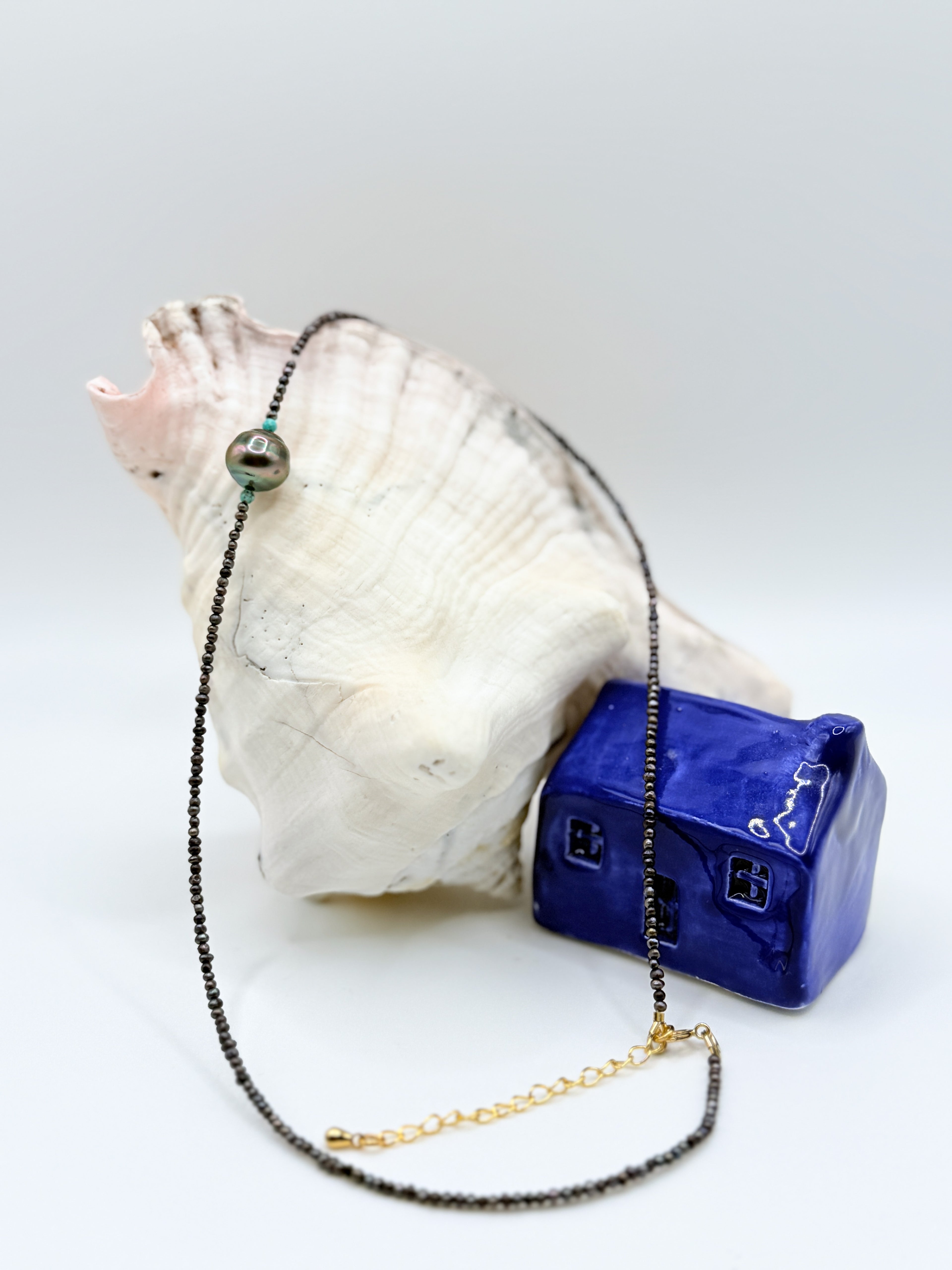 Delicate choker necklace with Tahitian baroque pearl centrepiece and tiny seed pearl beads draped over white conch shell next to blue ceramic house decoration